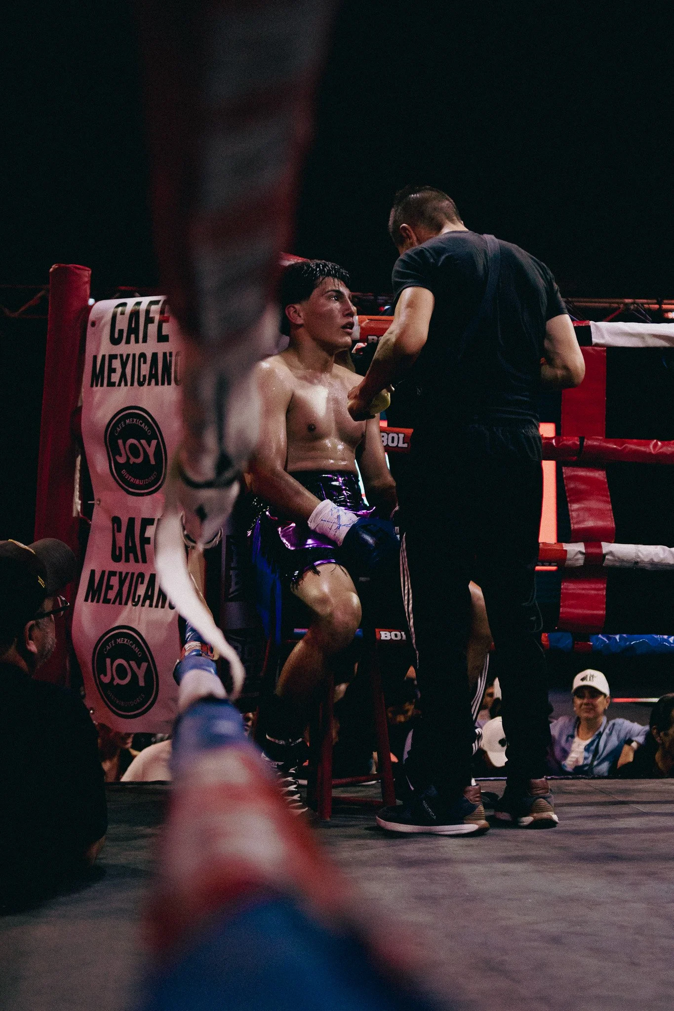 Cuerpo de un boxeador sentado en la esquina del ring, recibiendo instrucciones de un entrenador en medio de un combate, con una expresión de cansancio y concentración, mientras una multitud lo observa.