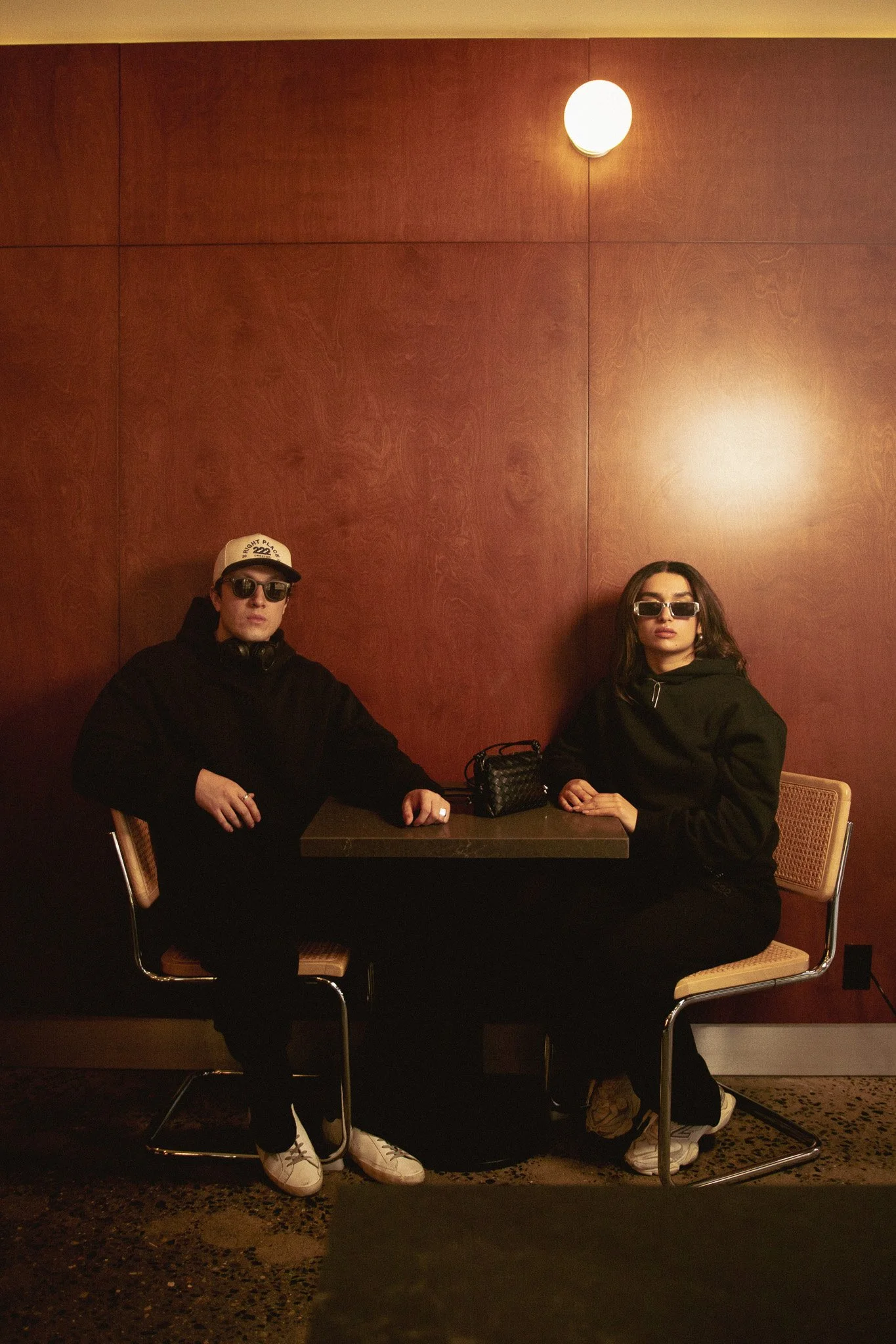 Dos personas sentadas en una mesa en un restaurante o cafetería, vestidas con ropa oscura y gafas de sol, con pared de madera de fondo y una luz circular en la pared.