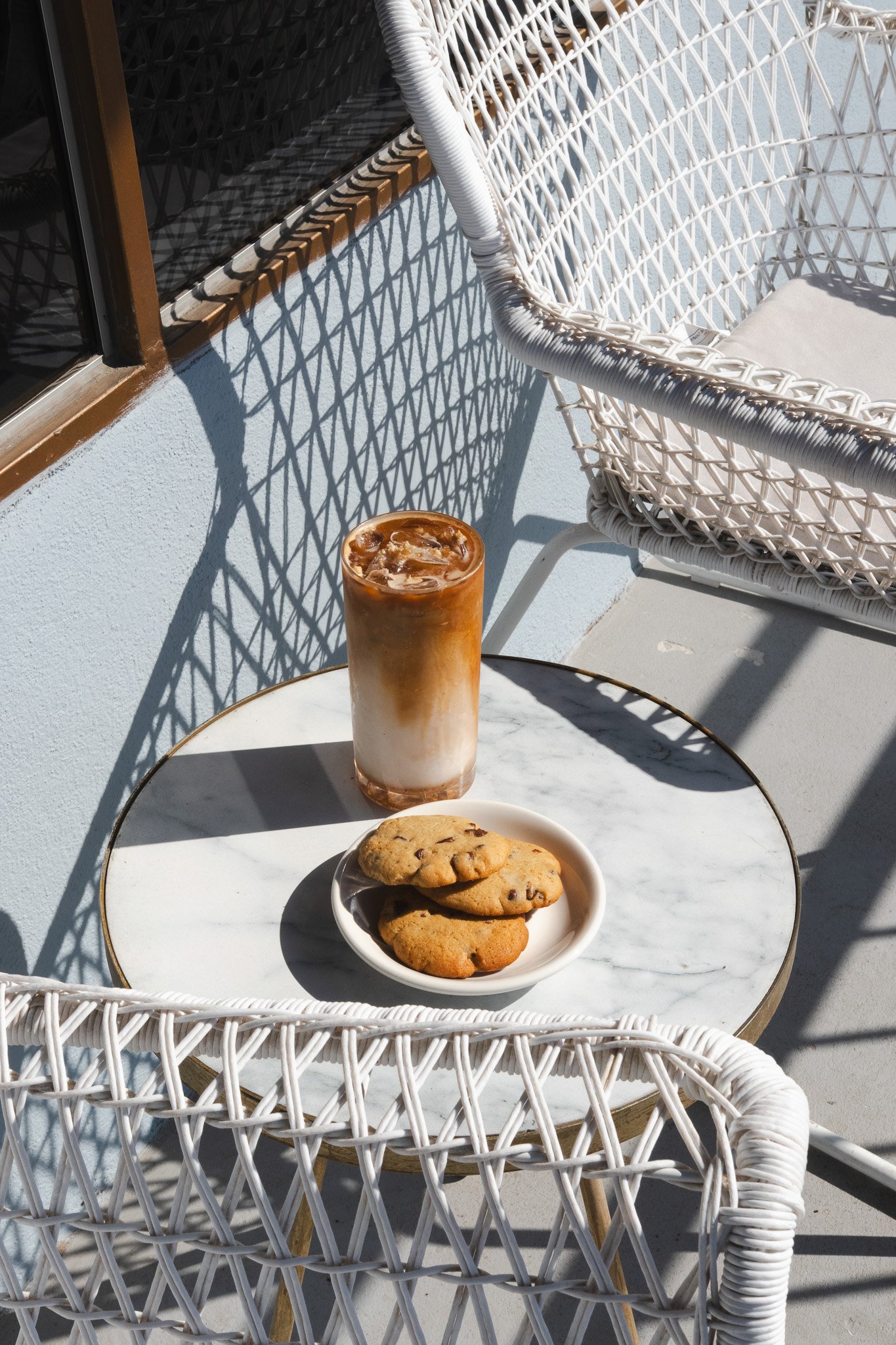 Una bebida de café con leche y hielo en un vaso alto, junto a tres galletas con chispas de chocolate en un plato pequeño sobre una mesa de mármol blanco, en una terraza con sillas de mimbre blancas y sombra del sol.