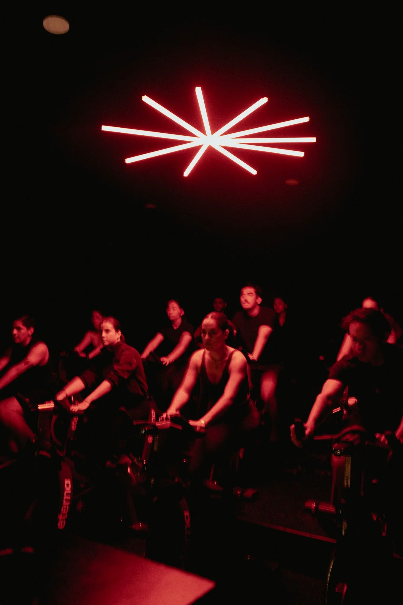 Varias personas haciendo ejercicio en una clase de ciclismo en interiores, en un entorno oscuro con luces rojas y un diseño de luces en forma de estrella en el techo.