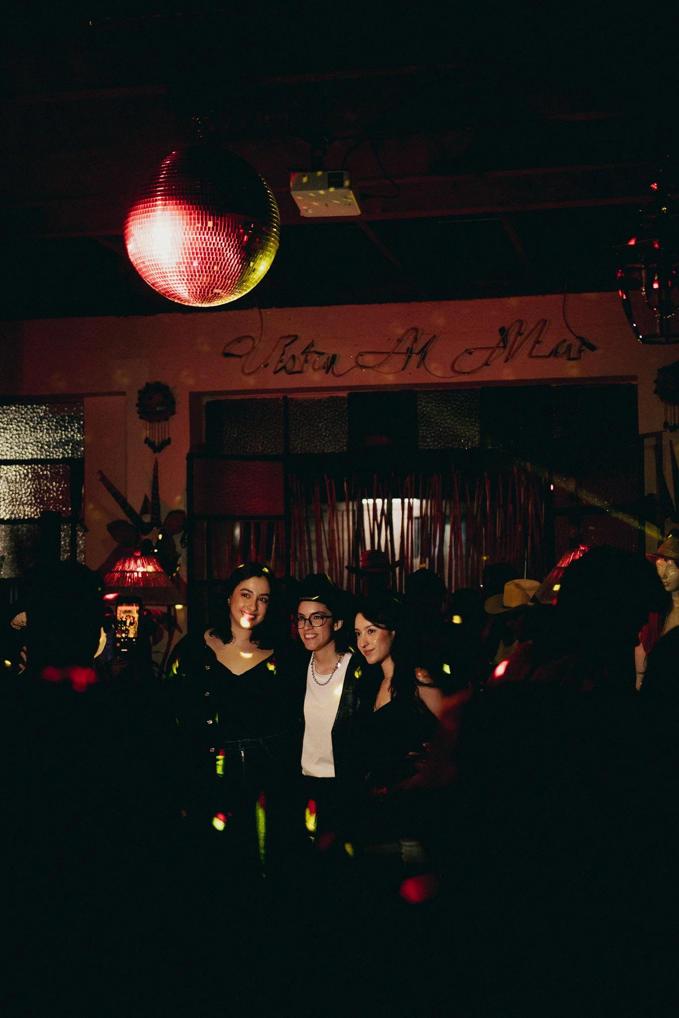 Grupo de personas en una fiesta en un lugar con decoraciones mexicanas y un globo de discoteca, con iluminación tenue y ambiente festivo.