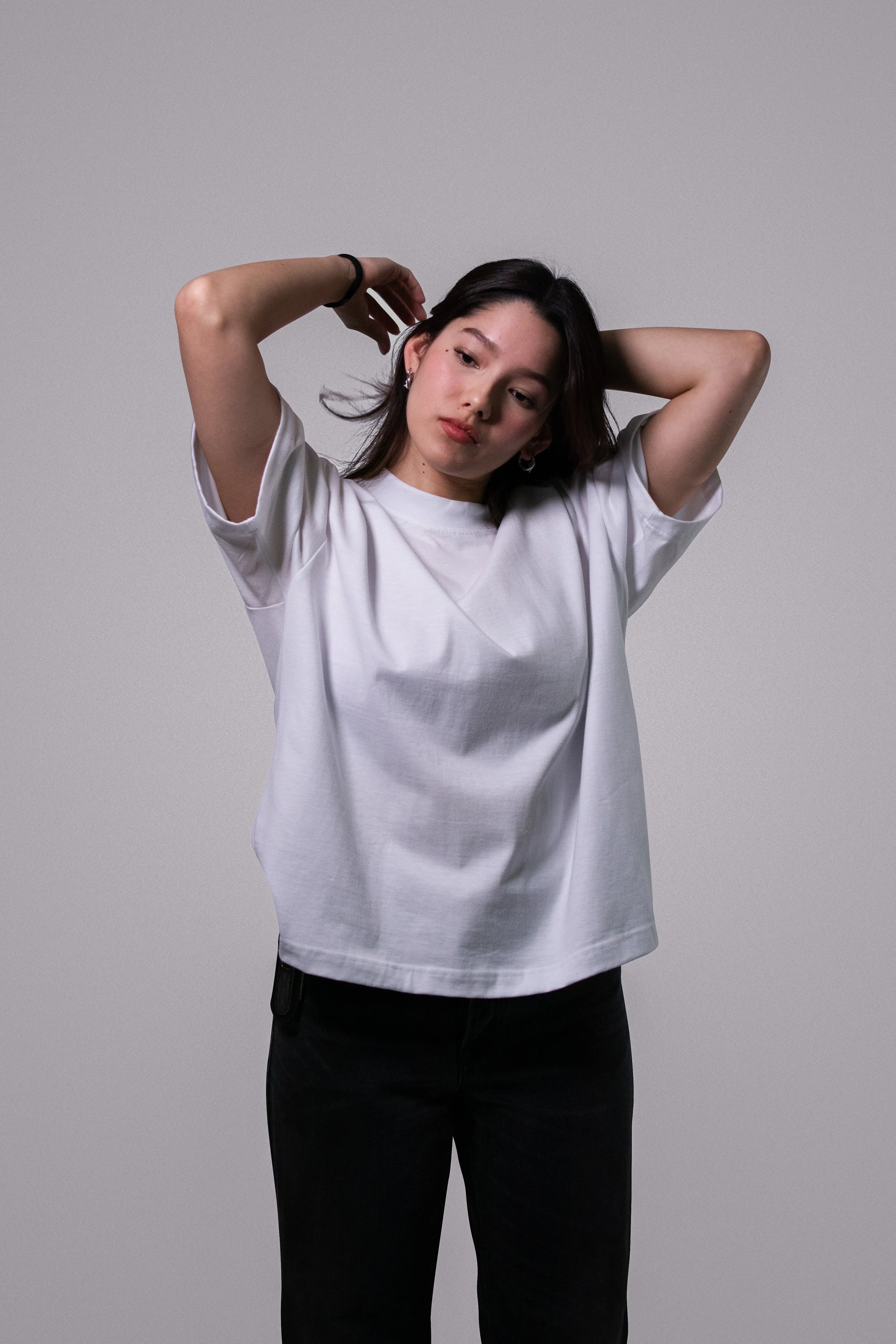 Joven mujer de cabello oscuro, con camiseta blanca y pantalones negros, posando con las manos en la cabeza, against a un fondo gris.