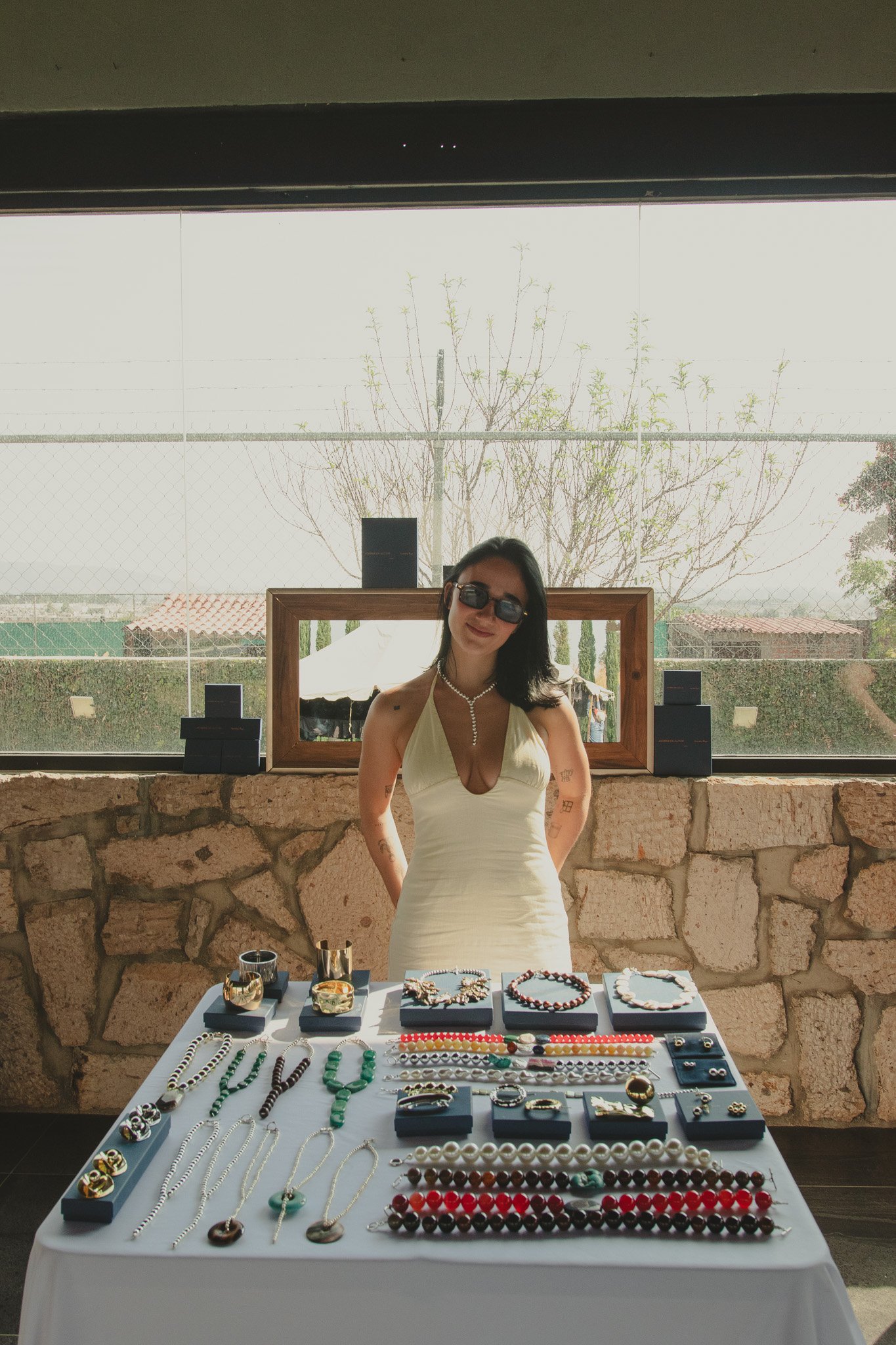 Mujer vendiendo joyería en mesa con collares y pulseras, con fondo de ventana y paisaje exterior.