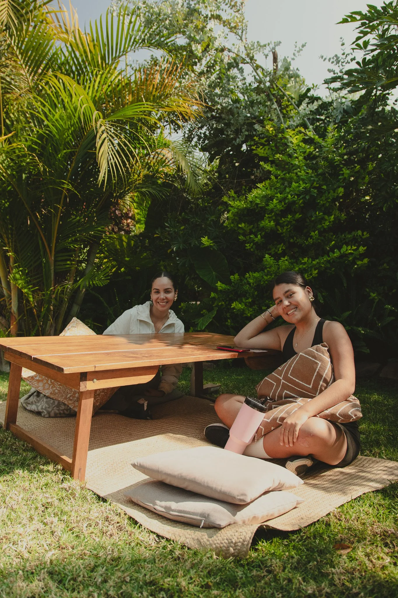 Dos jóvenes sonriendo en un ambiente al aire libre rodeados de plantas verdes, uno sentado en el suelo con cojines y el otro apoyado en una mesa de madera.