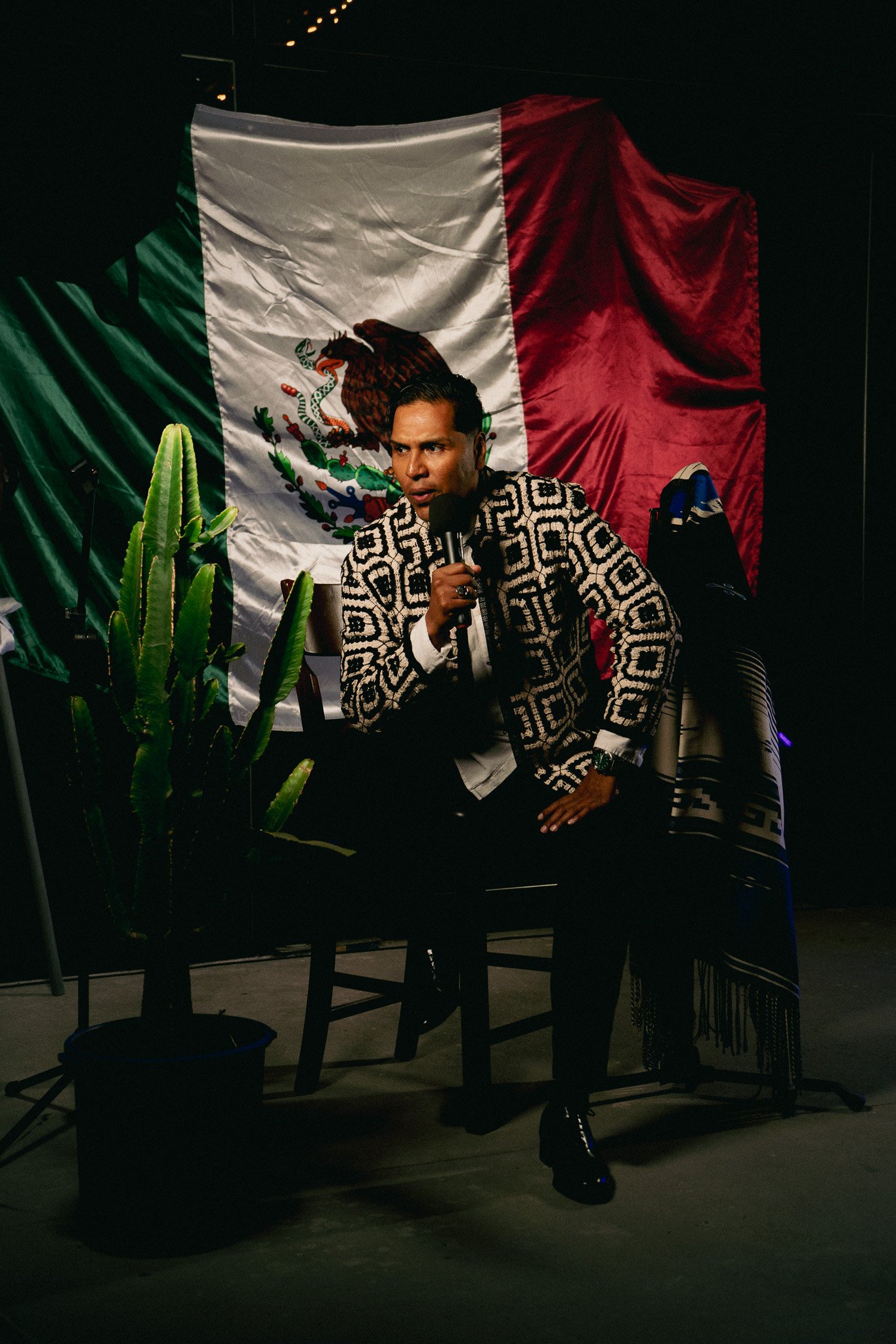 Hombre sentado en una silla con un micrófono en la mano, frente a una bandera de México y decoraciones culturales, iluminado en un entorno oscuro.