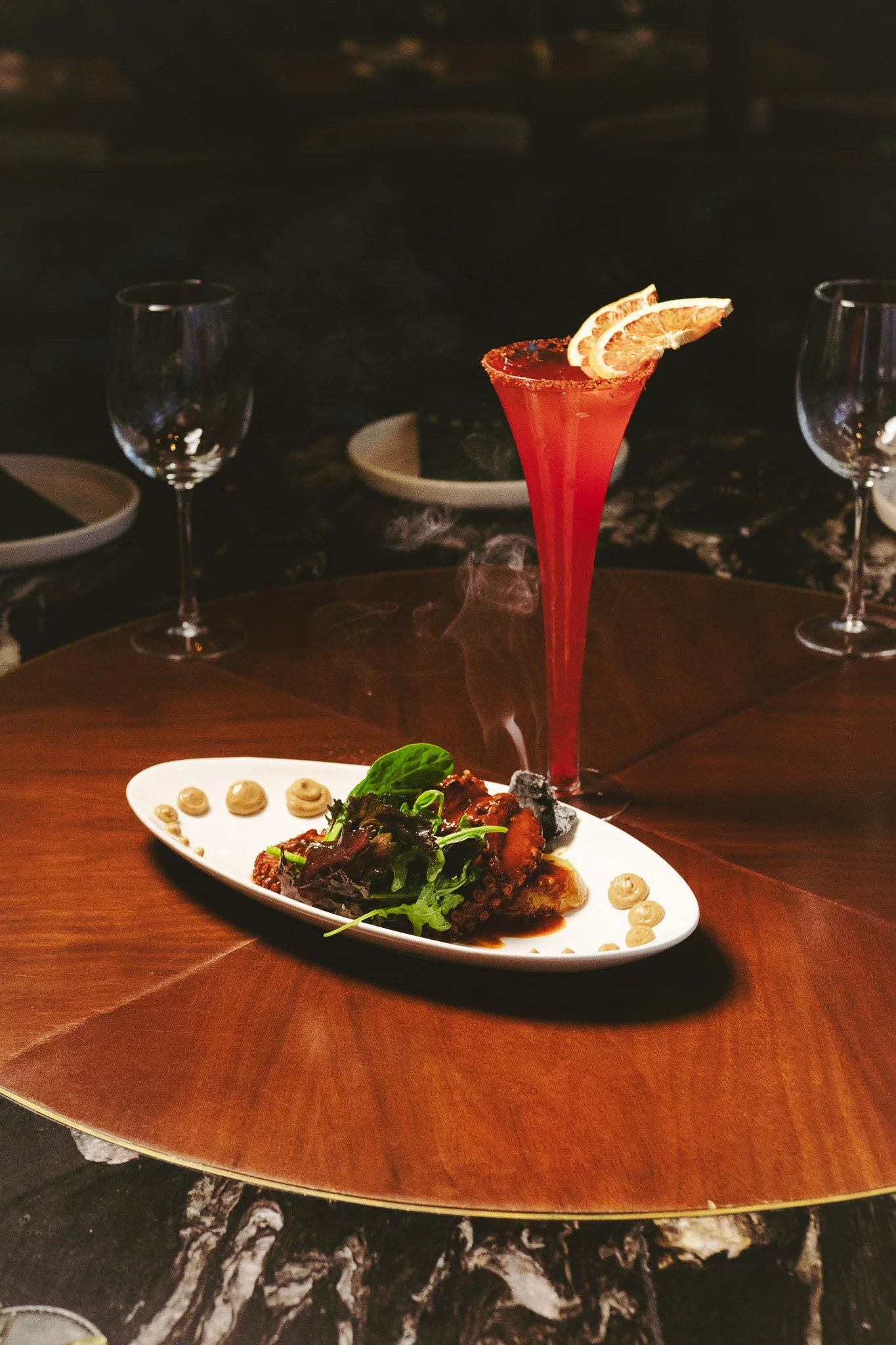 Cóctel rojo con cáscara de limón en una copa alta y plato con mariscos y ensalada en una mesa de restaurante.