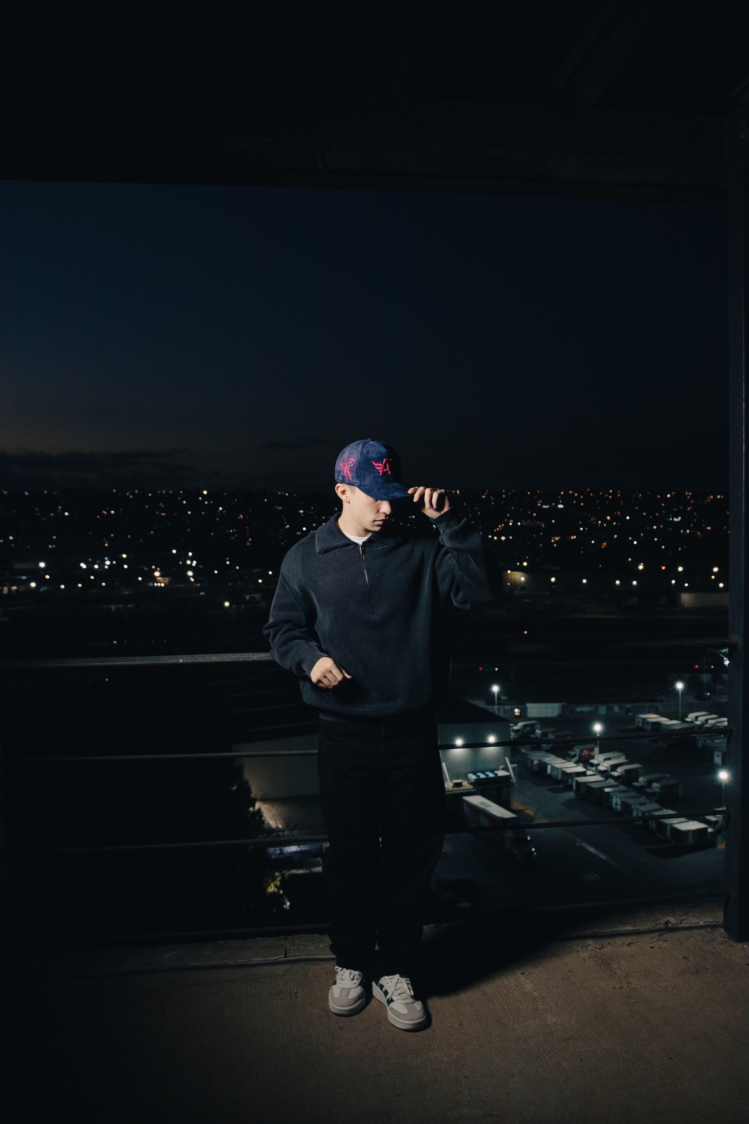 Joven con sombrero y sudadera de pie en un balcón con vista nocturna a la ciudad con muchas luces y edificios, sosteniendo la visera del sombrero con la mano.
