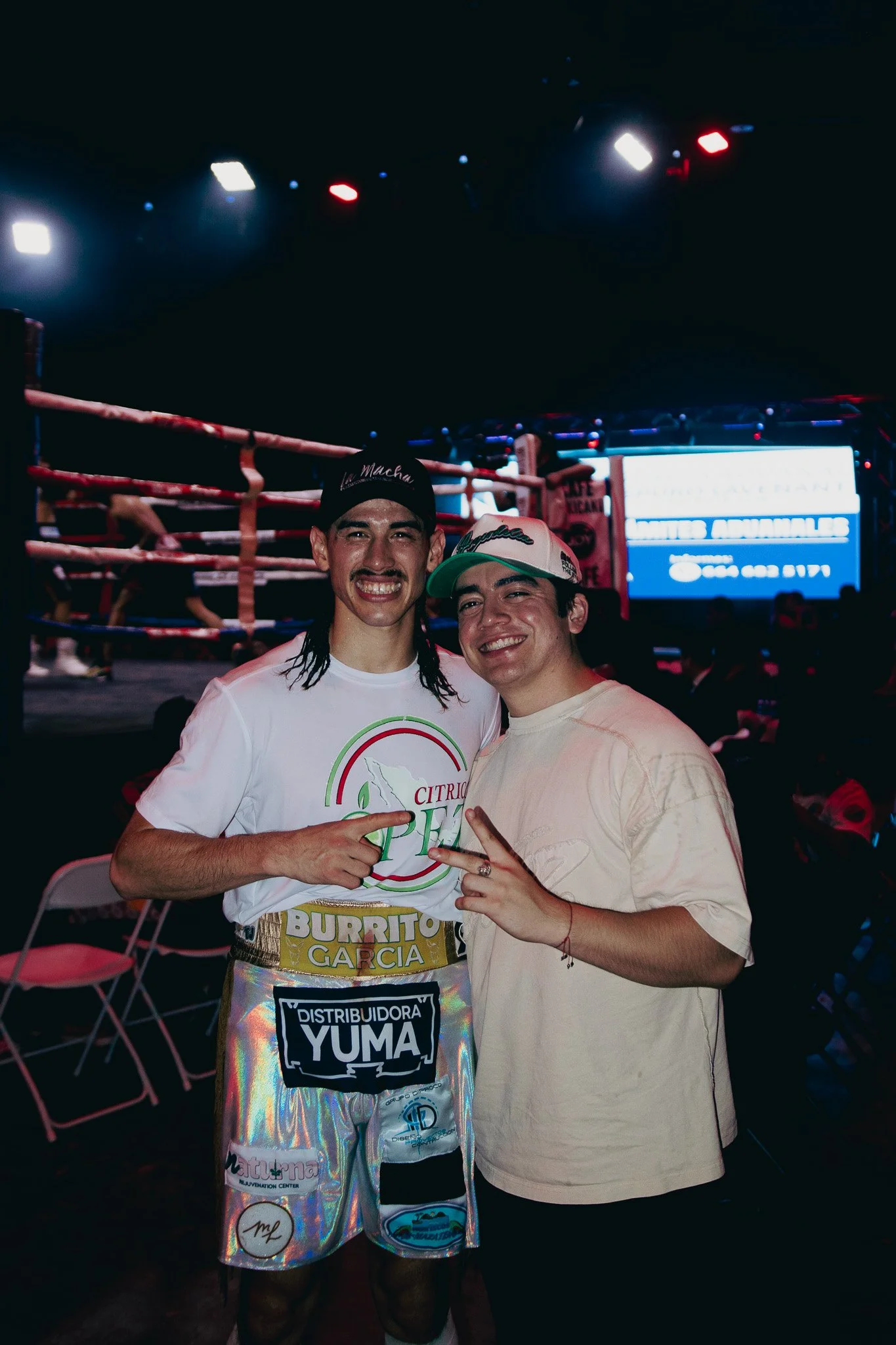 Dos hombres en un evento de boxeo, con ropa de boxeador, en un ring, sonriendo y señalando con las manos. Tras ellos, sillas y una pantalla iluminada.