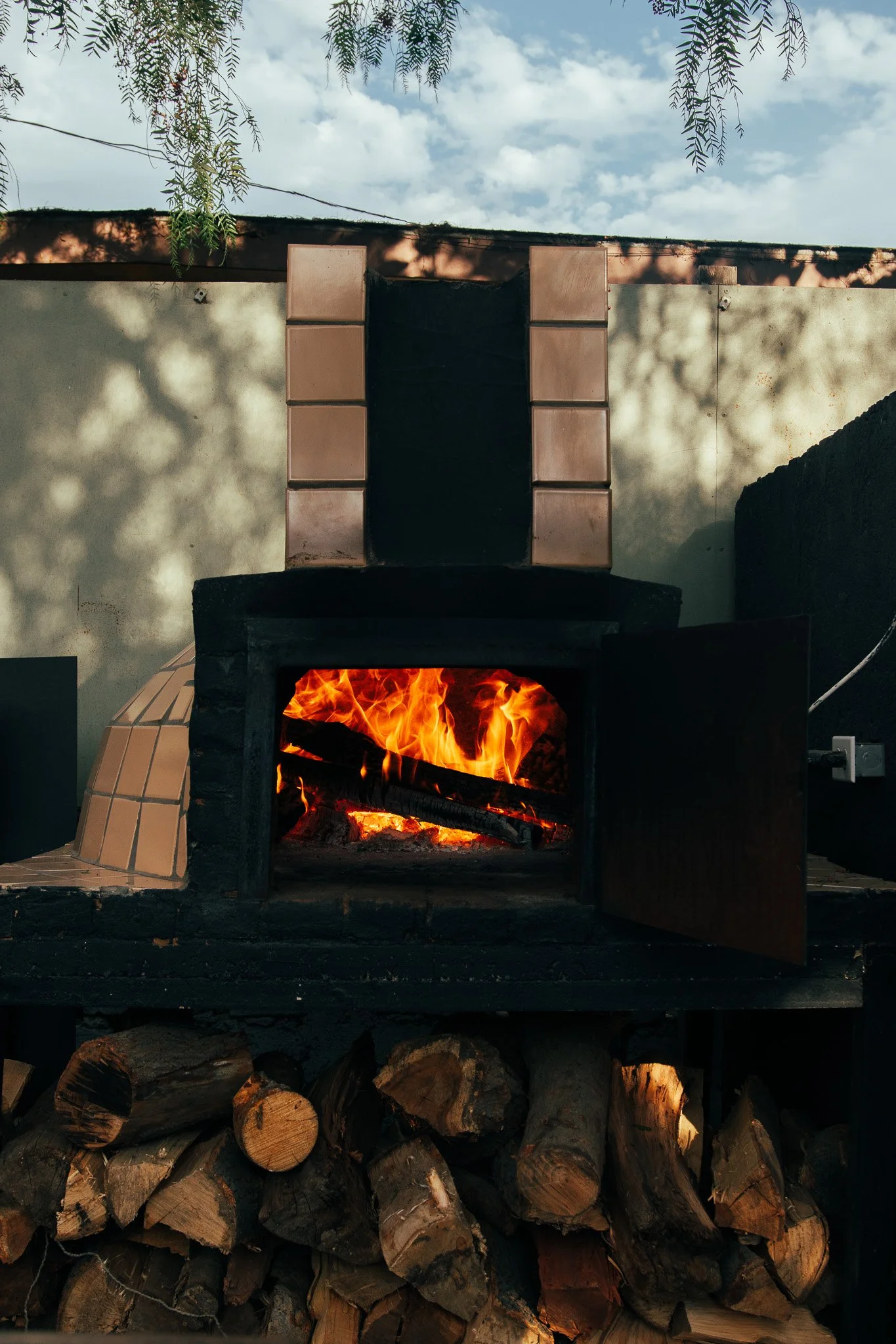 Horno de leña encendido con fuego visible, apilado de madera debajo, y fondo de muro con sombra de ramas y cielo.