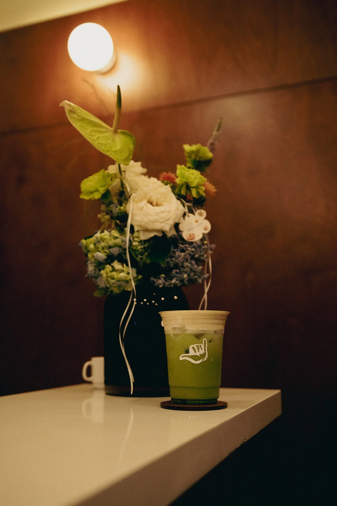 Vaso con una bebida verde sobre una mesa, al fondo un jarrón con flores y una luz en la pared.