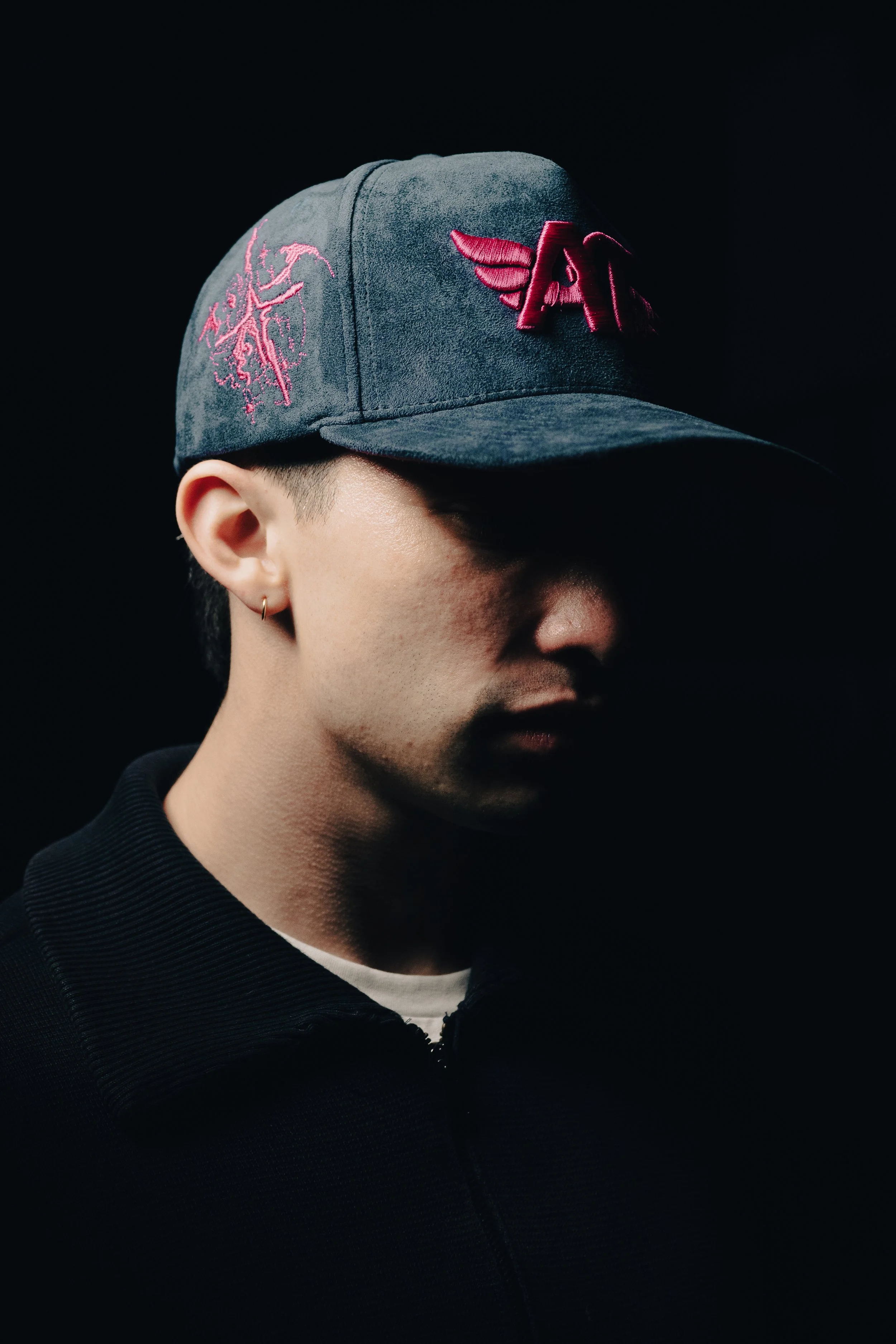 Hombre con gorra negra con logotipo de ala en la parte frontal, en un fondo negro. Solo parte de su rostro y oreja son visibles.