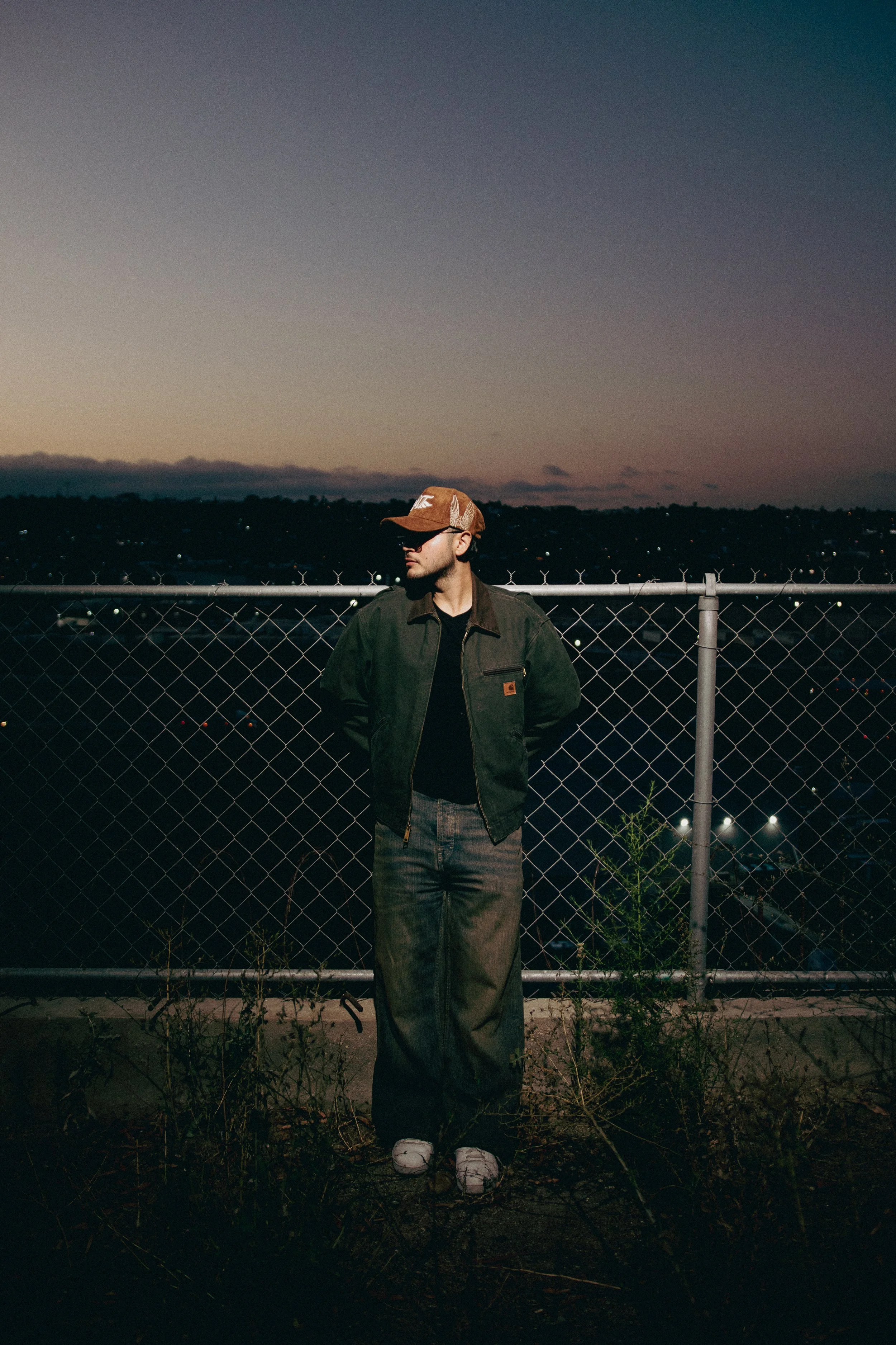 Joven con gorra y lentes de sol, de pie junto a una cerca metálica, en una ciudad al atardecer con un cielo nublado y luces en el fondo.