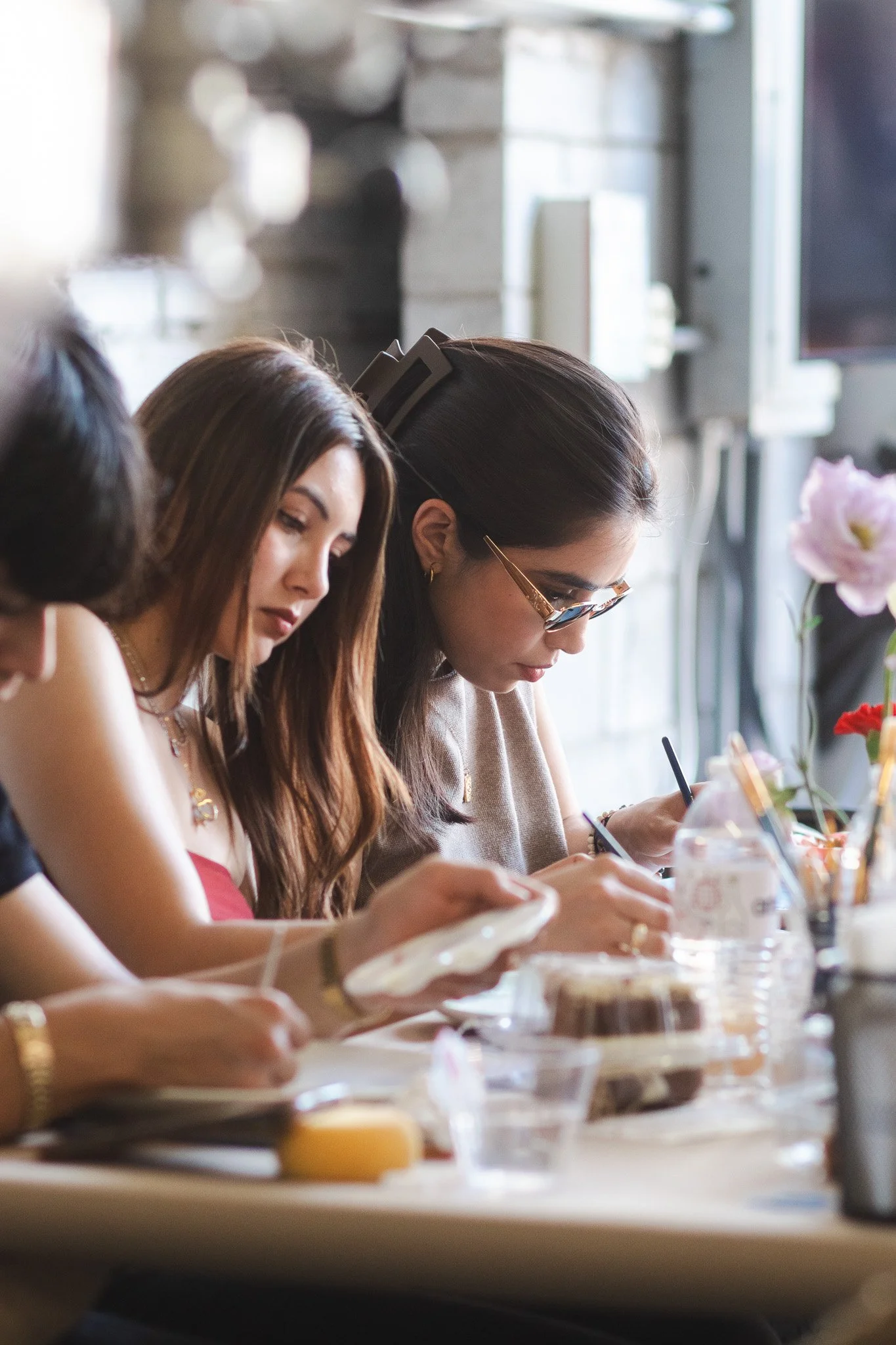 Mujeres participando en actividad artística con herramientas y materiales en una mesa, en un espacio interior con luz natural.