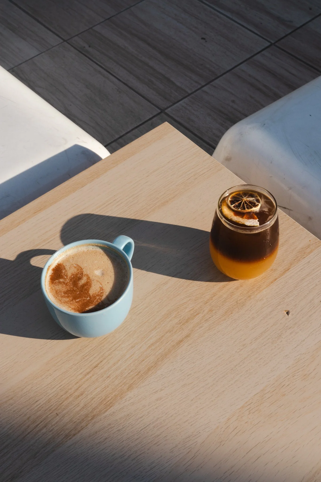 Una taza azul con café con leche y una bebida en un vaso con rodaja de limón en hielo, sobre una mesa de madera clara.
