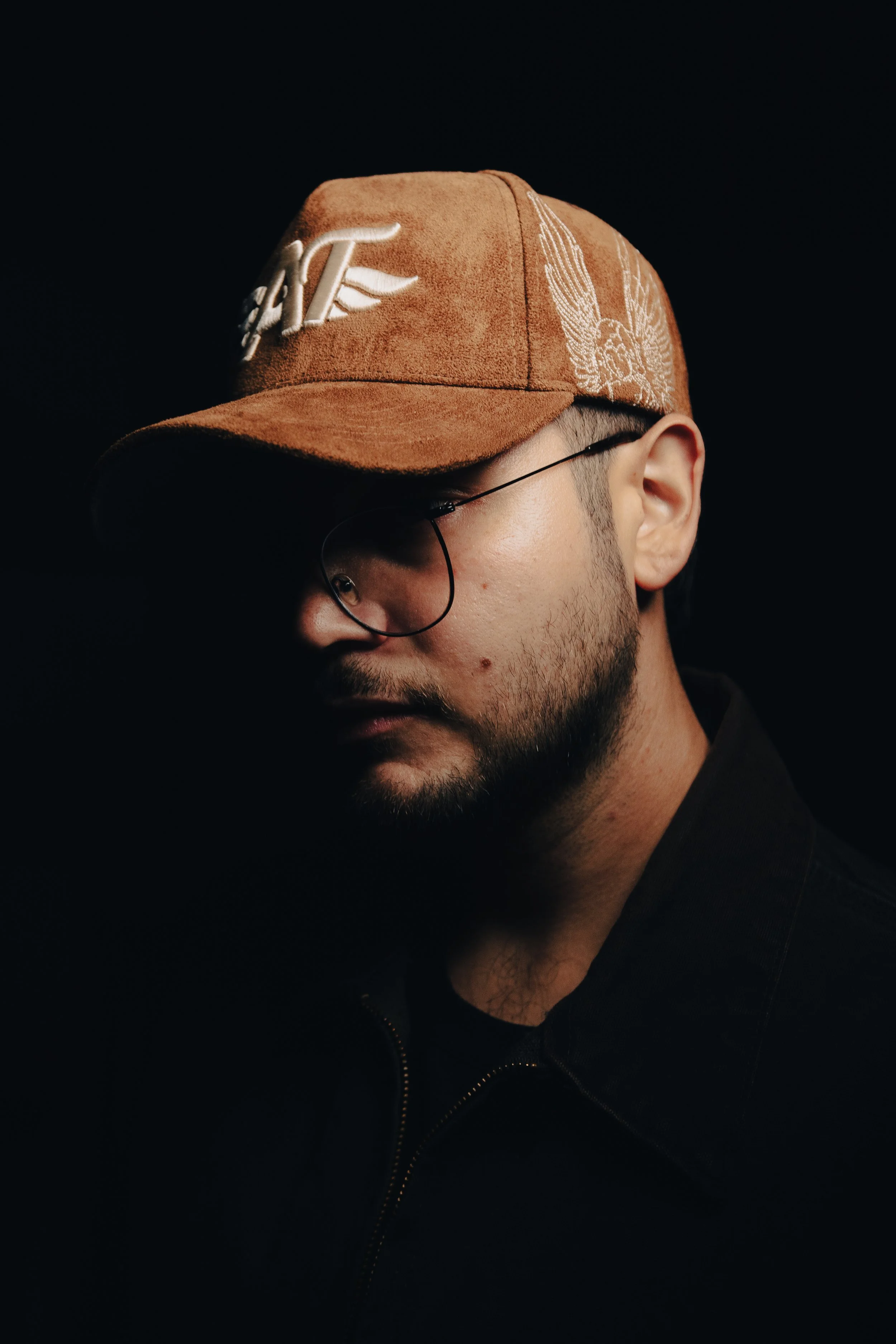 Hombre con barba, gafas y gorra marrón con logotipo y diseño de alas, usa chaqueta negra y está en fondo oscuro.