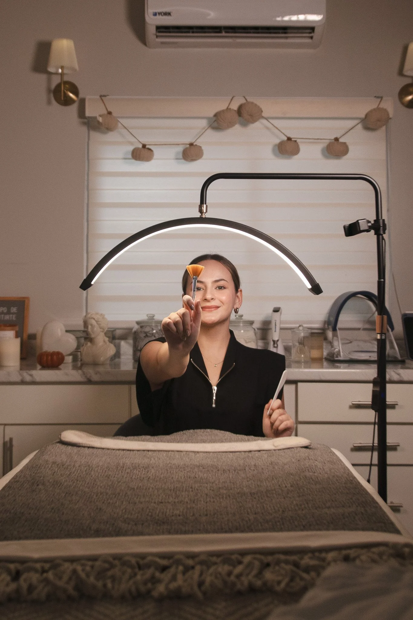 Mujer en un salón, sosteniendo una brocha y una tarjeta, bajo una lámpara circular, en un ambiente decorado con adornos en la pared.