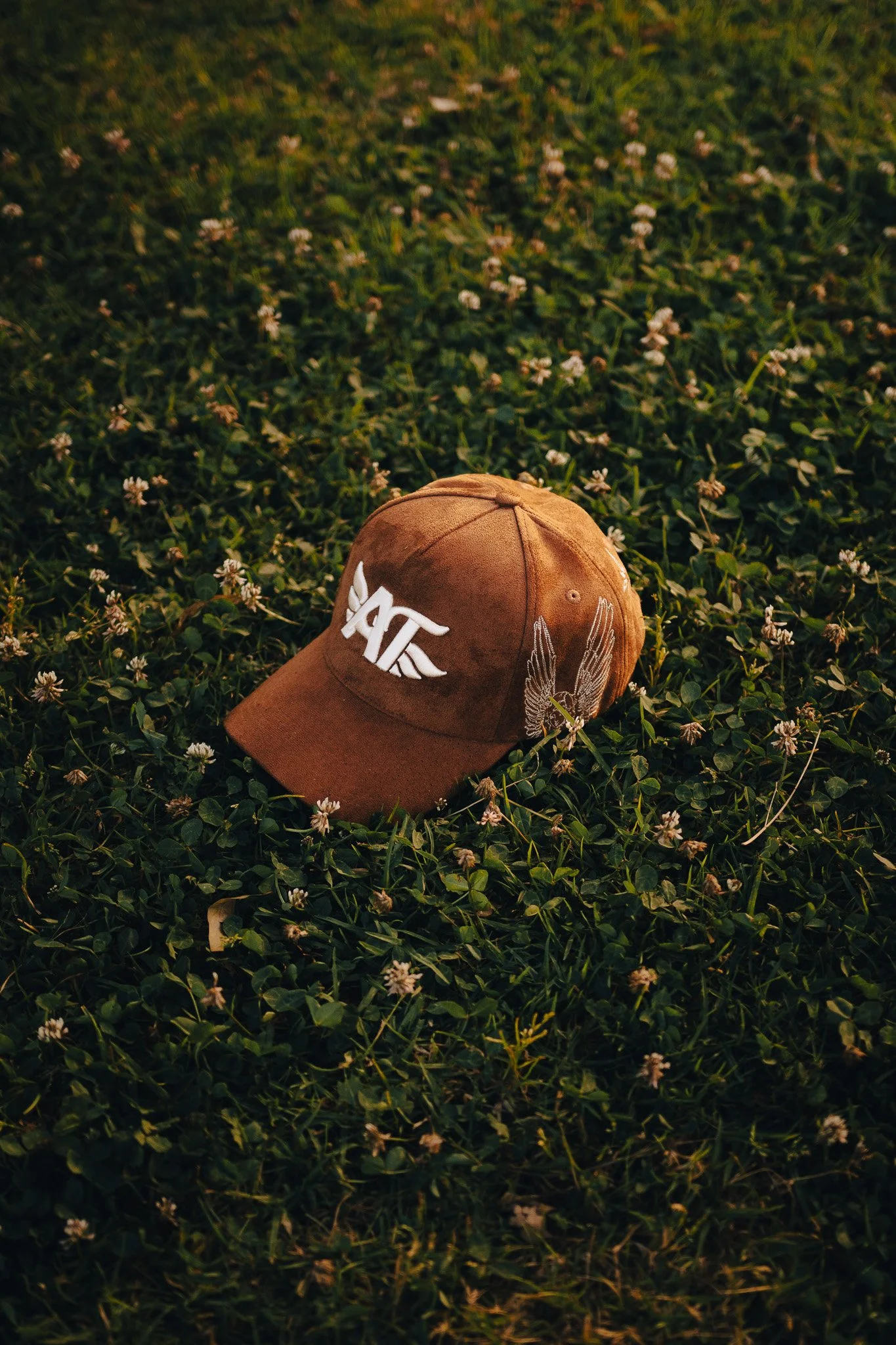 Gorra de color marrón con un diseño de alas y letras blancas, colocada sobre césped con pequeñas flores blancas.