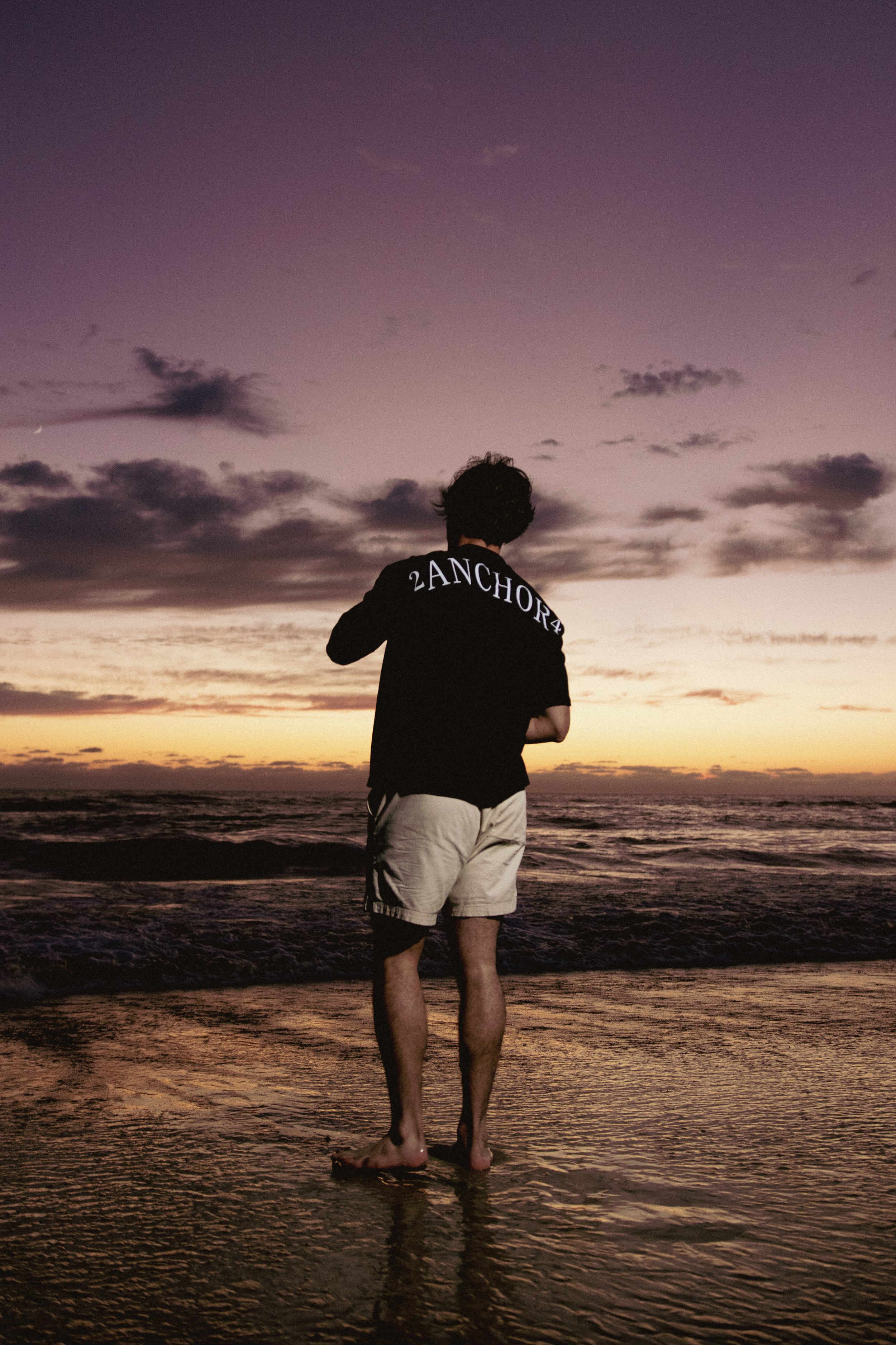 Joven con pantalones cortos y camiseta gráfica en la orilla del mar durante el atardecer.