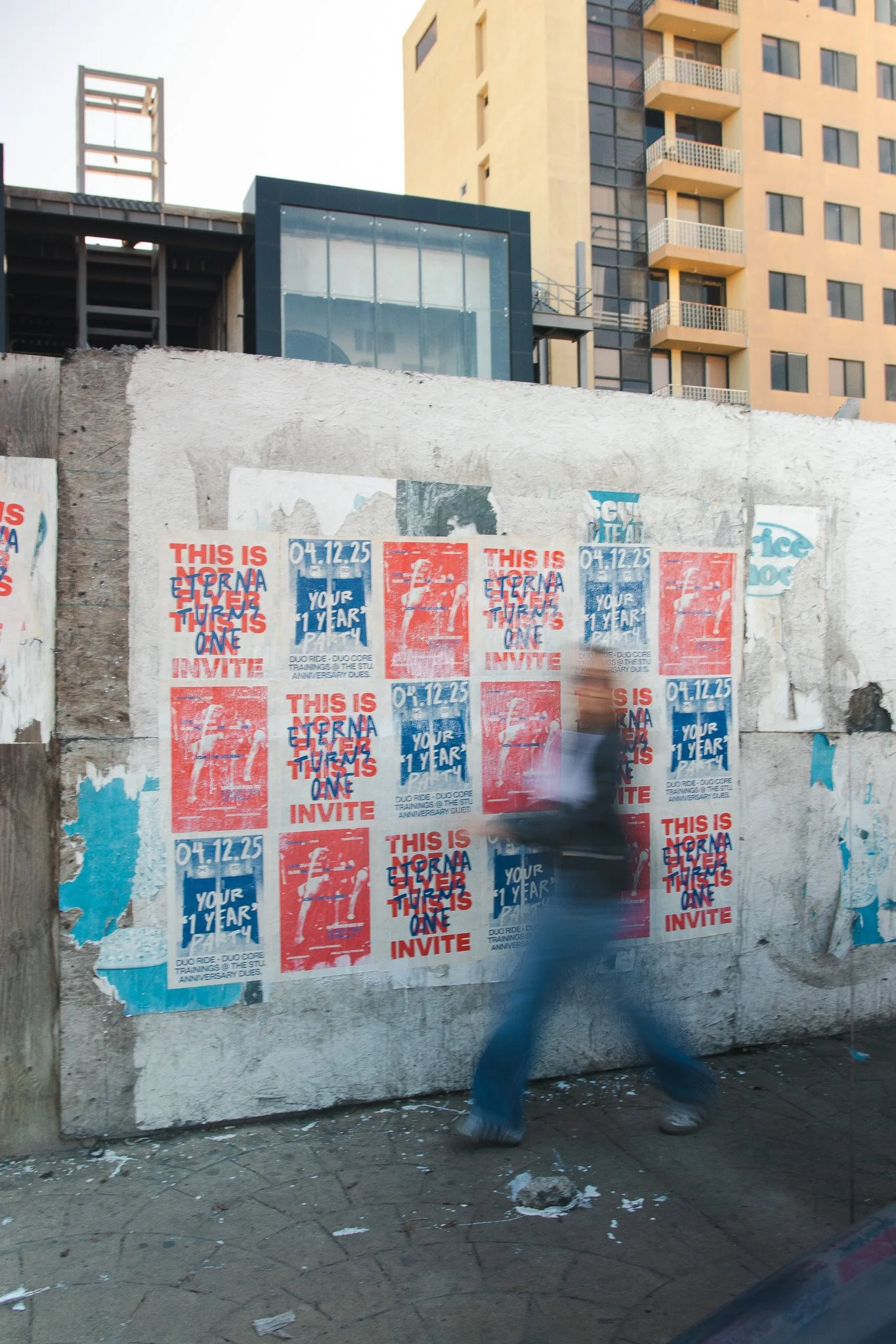 Mural con carteles publicitarios en la calle, en un entorno urbano con edificios altos al fondo, y una persona caminando rápidamente frente a la pared.