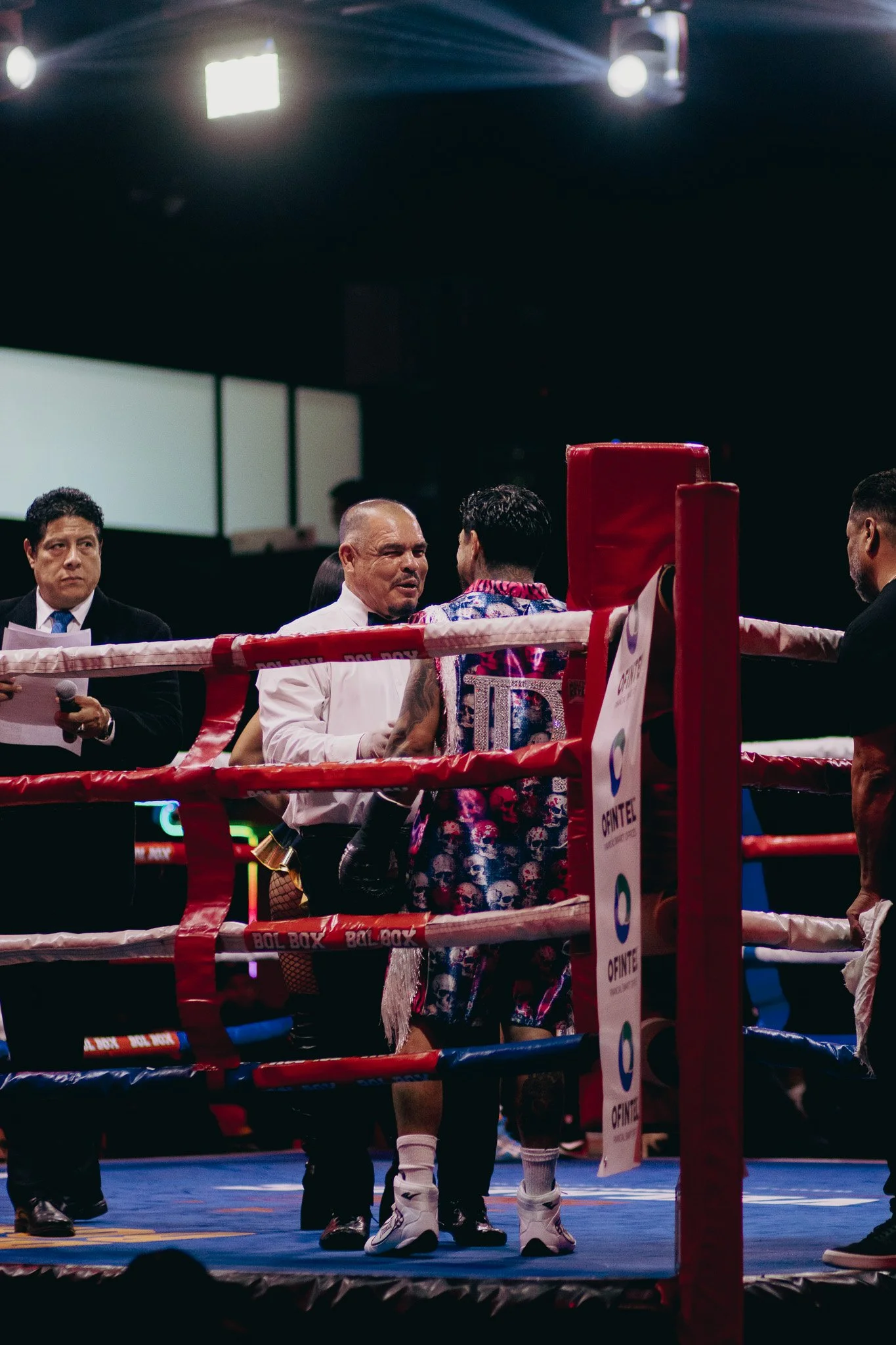 Hombres en un ring de boxeo discuten durante un evento de boxeo, uno lleva un chaleco con dibujos de calaveras y el otro viste una camisa con estampado colorido.