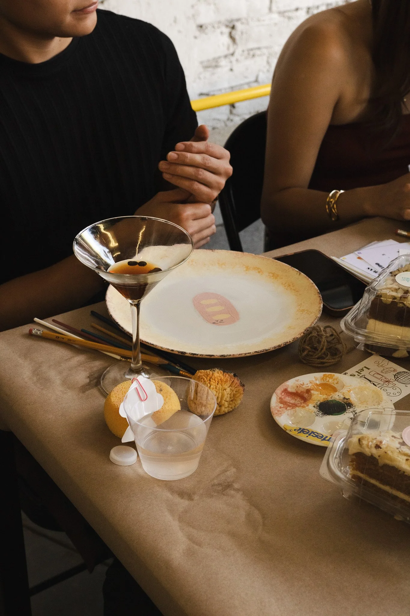 Mesa con platos vacíos y restos de comida, un cóctel en copa de cóctel y una bebida en vaso plástico, junto a utensilios, postres y otros objetos, en un ambiente informal.