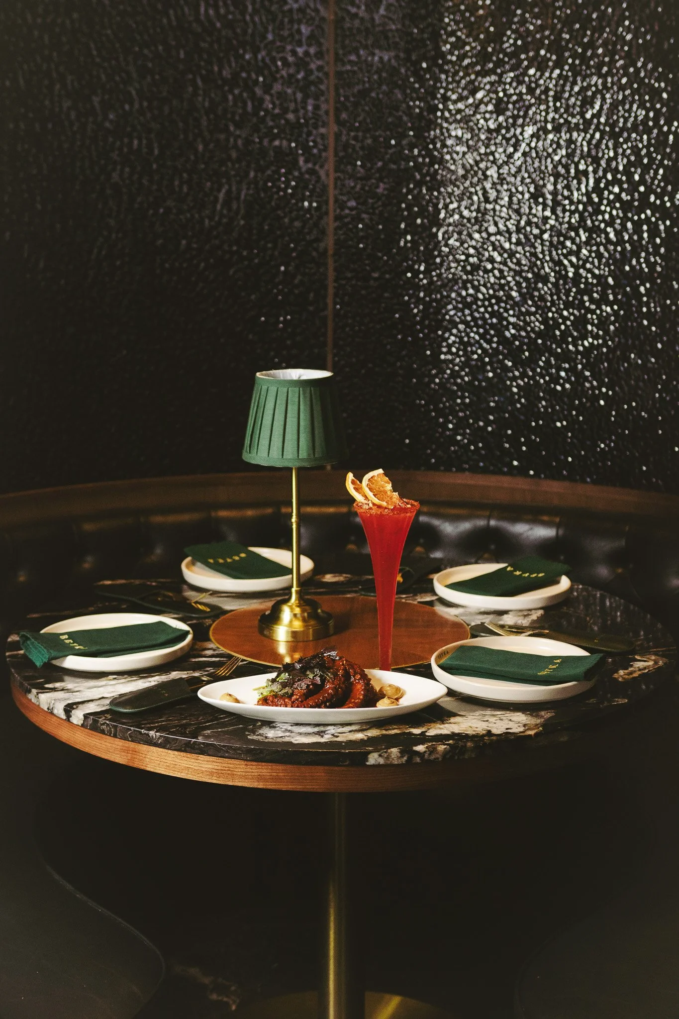 Mesa elegant con cinco platos, una lámpara de mesa verde, fondo negro con textura y palabras en oro en los cubiertos.