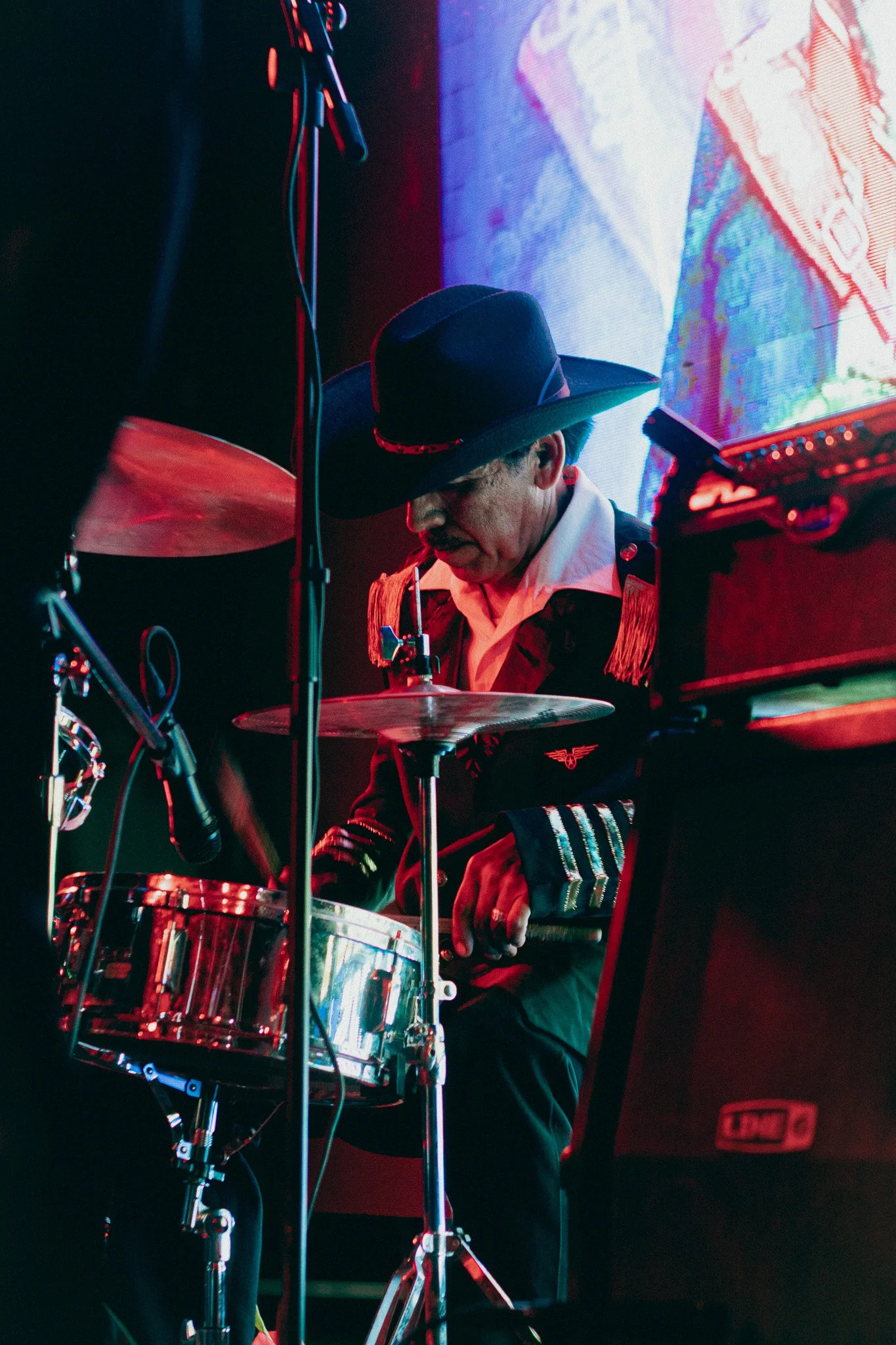 Un músico con sombrero negro, tocando la batería durante una actuación en un escenario con iluminación roja y púrpura.