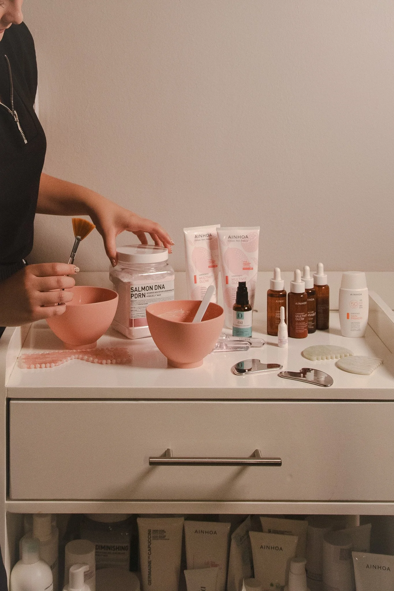 Productos de cuidado personal y cosmética en una mesa, incluyendo frascos, tubos y herramientas como espejos y esponjas, con una persona que sostiene un frasco y una brocha.