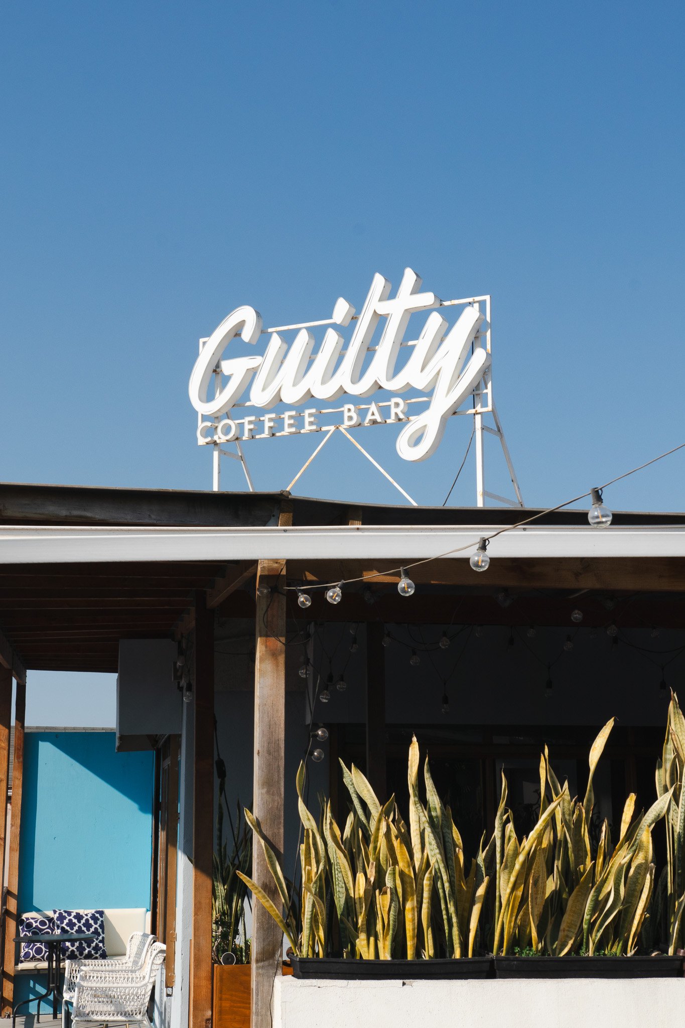 Señal luminosa que dice 'Guilty Coffee Bar' en un cielo azul, con plantas altas en el primer plano y luces decorativas colgadas.