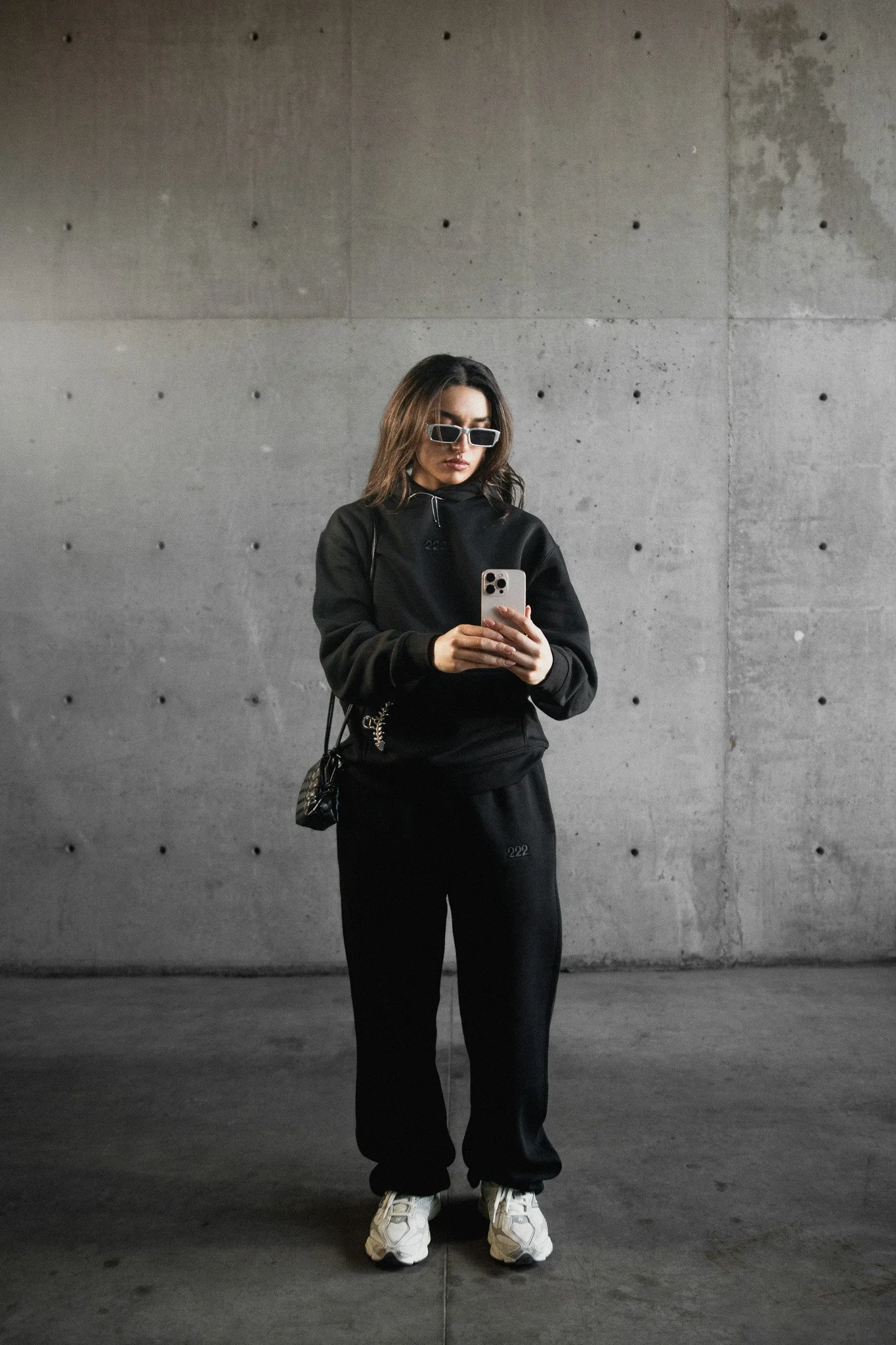 Joven mujer con gafas de sol, ropa deportiva negra, bolso pequeño, tomando selfie con teléfono en un espacio con paredes de concreto