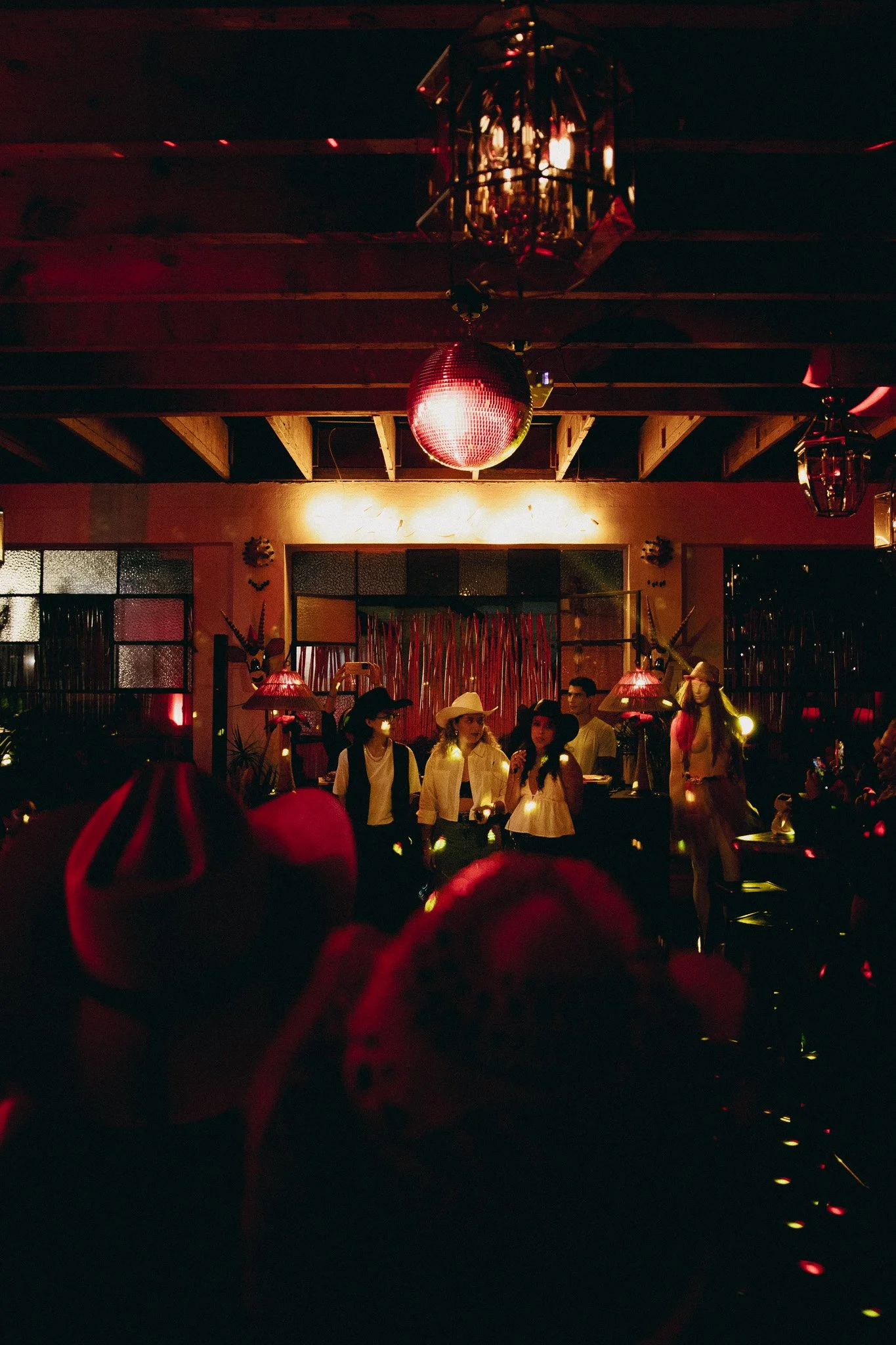 Escena en un club nocturno con decoraciones de estilo mexicano, como sombreros y máscaras, con varias personas socializando y disfrutando del ambiente, iluminada con luces de colores y focos suaves.