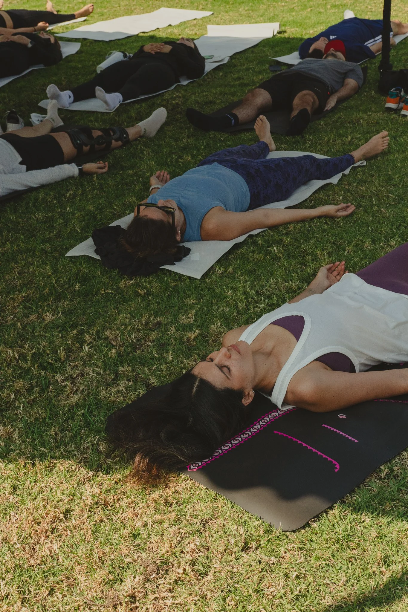 Varias personas en ropa deportiva realizan yoga o meditación en un campo de césped, acostadas en colchonetas y en posición de relajación.