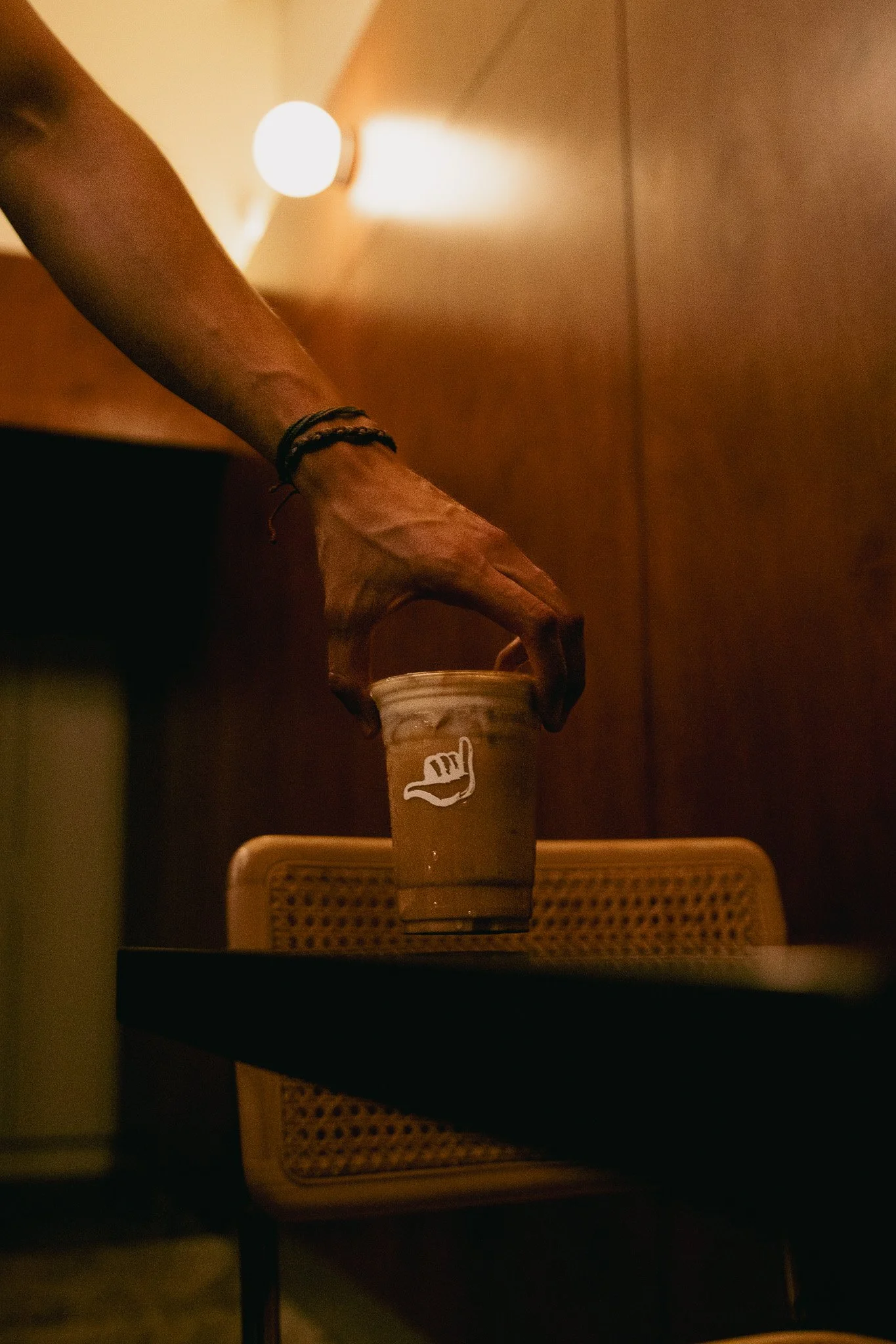 Una mano colocando una taza de café en una mesa, con un muro de madera y luz tenue en el fondo.