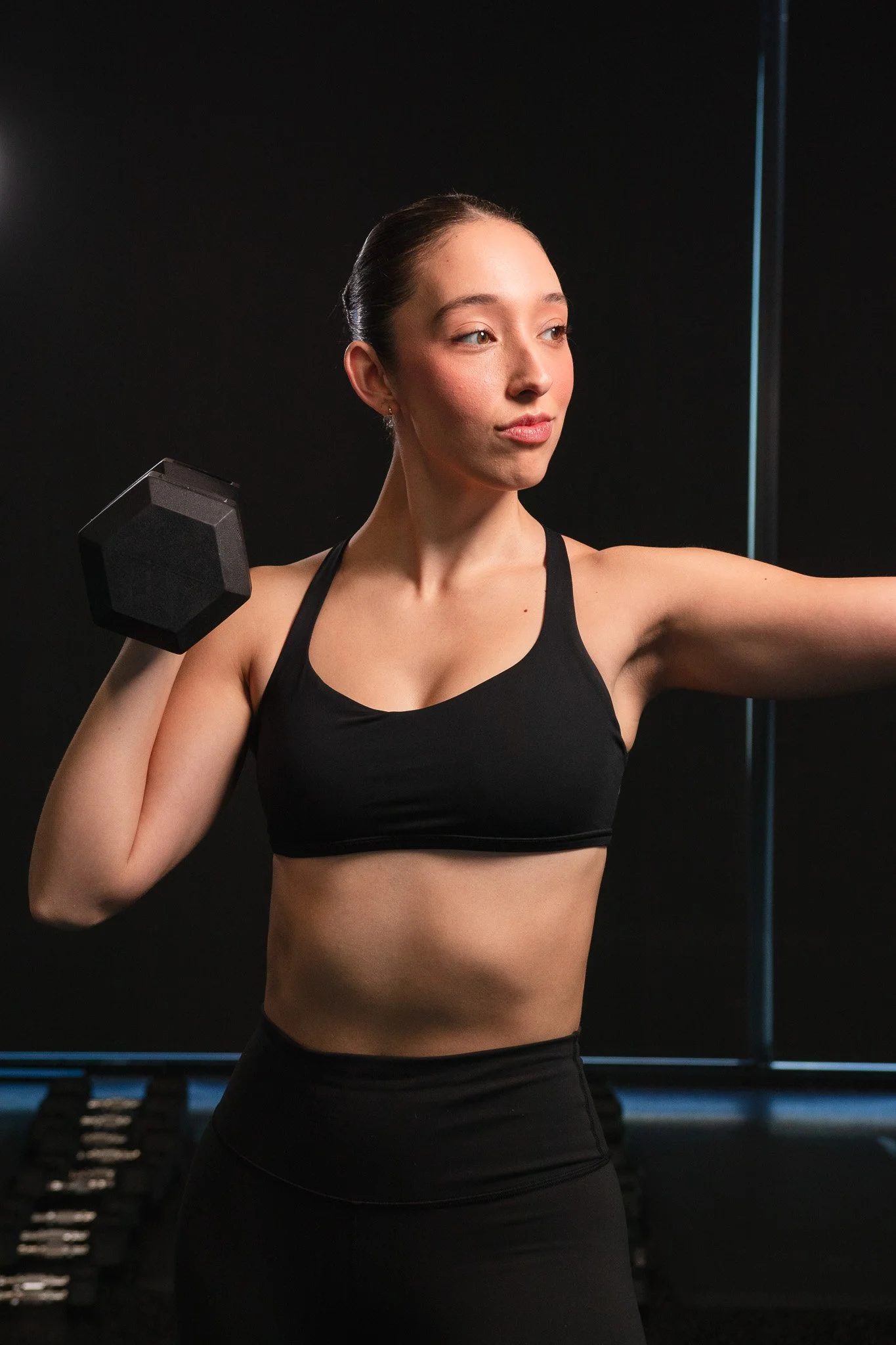 Mujer levantando un peso en un gimnasio, vestida con ropa deportiva negra, en un entorno de entrenamiento.