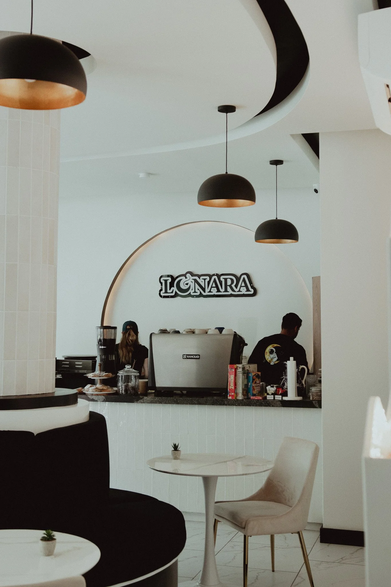 Interiores de una cafetería llamada "LÑNARA" con mostrador, dos empleados, lámparas negras, mesas blancas y sillas beige.