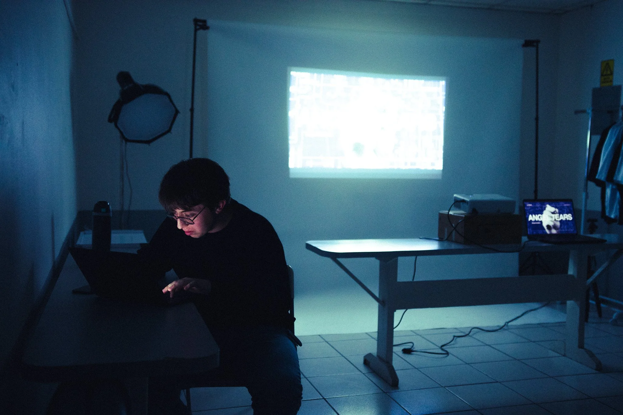Joven sentado en una mesa con una computadora portátil encendida, en una habitación oscura con proyector y pantalla que muestra contenido en la pared.