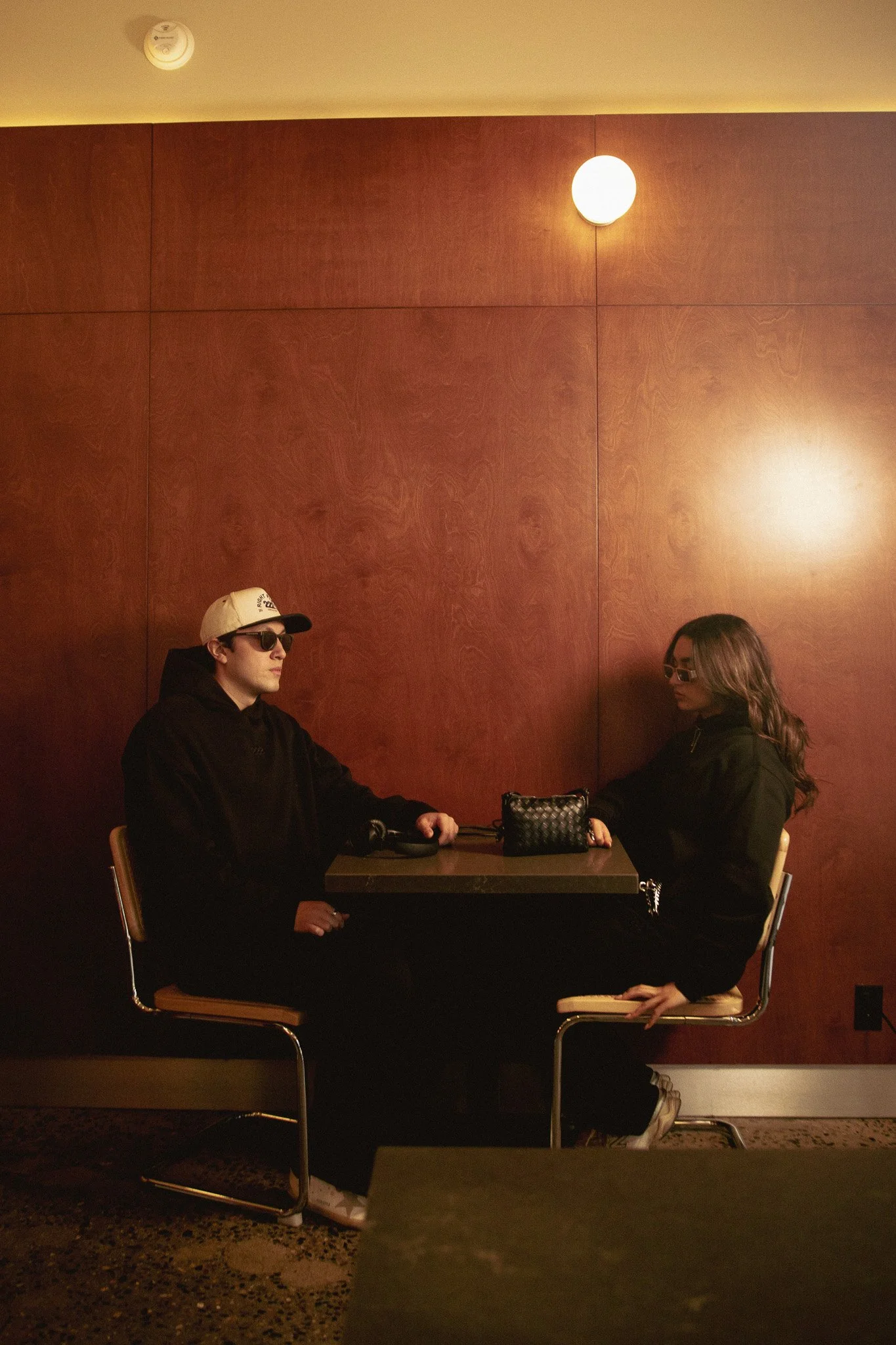 Dos personas sentadas en una mesa en un ambiente con paredes de madera, una mujer con gafas de sol y un hombre con gorra y gafas oscuras, parecen estar en conversación.