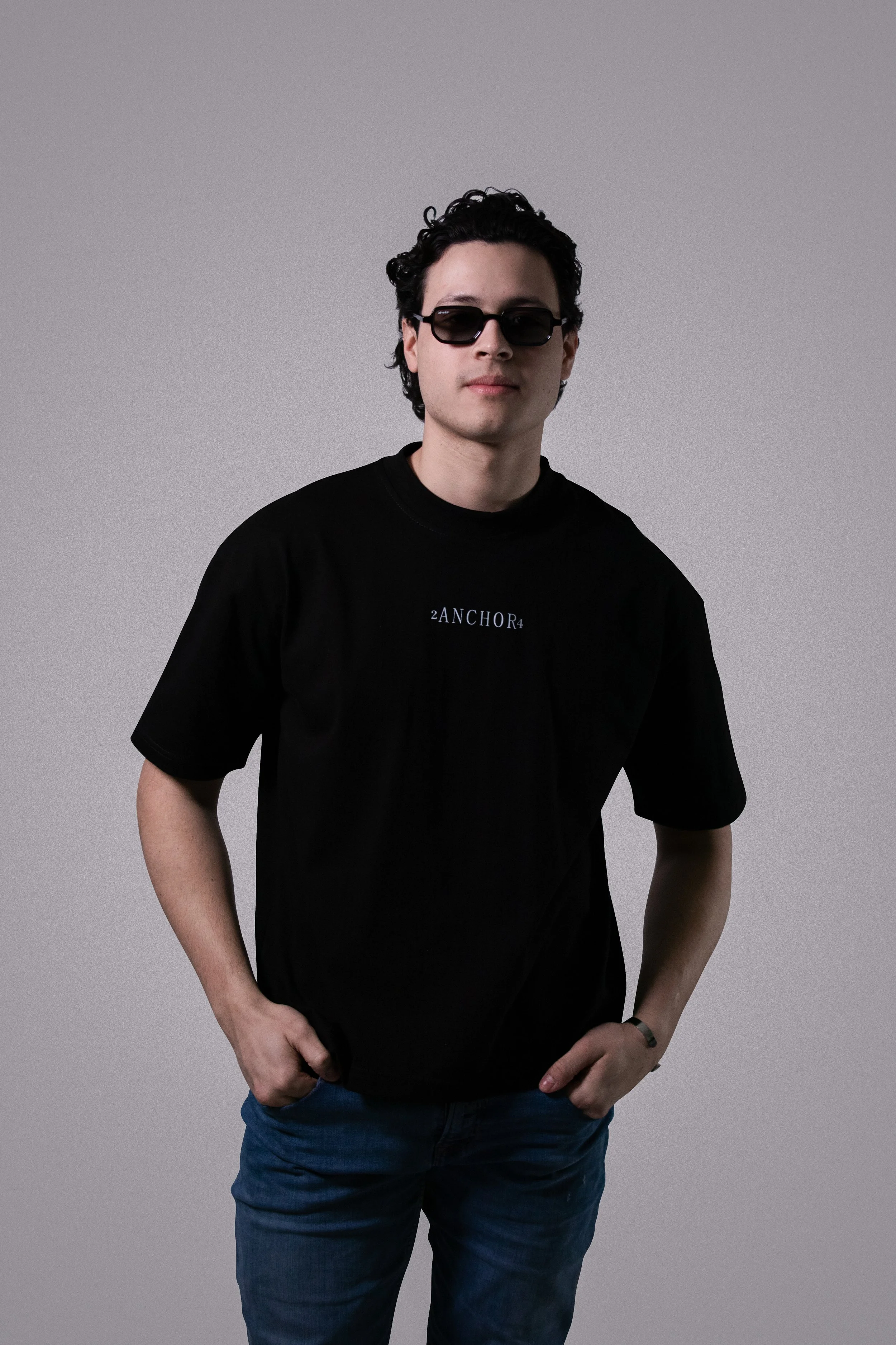 Joven con cabello rizado, gafas de sol negras, camiseta negra con texto en inglés y pantalones oscuros, de pie contra fondo gris liso.