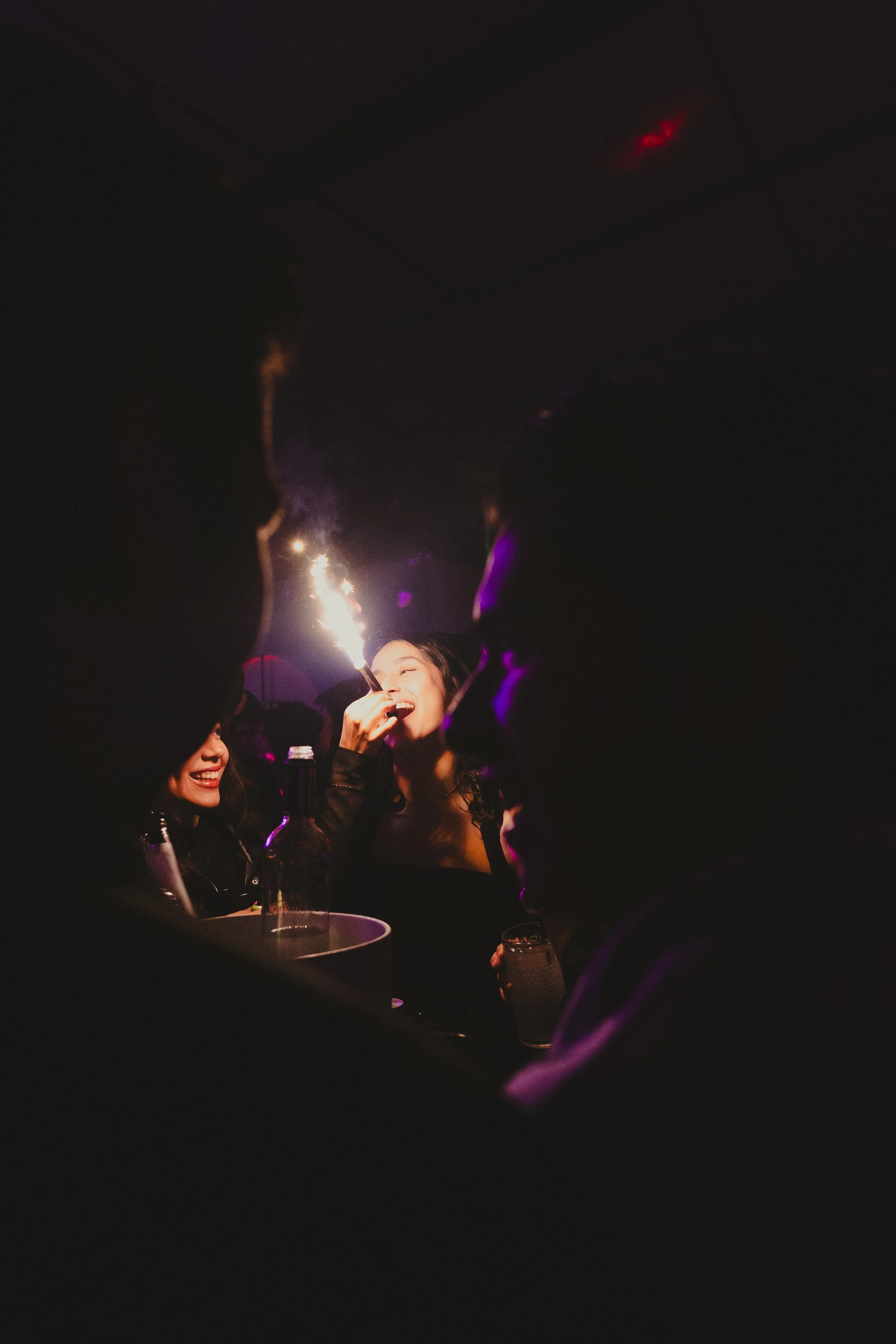 Personas en celebración en un espacio oscuro con fuegos artificiales, una mujer sonriente sosteniendo un fósforo o vela, y otras personas en el fondo disfrutando.