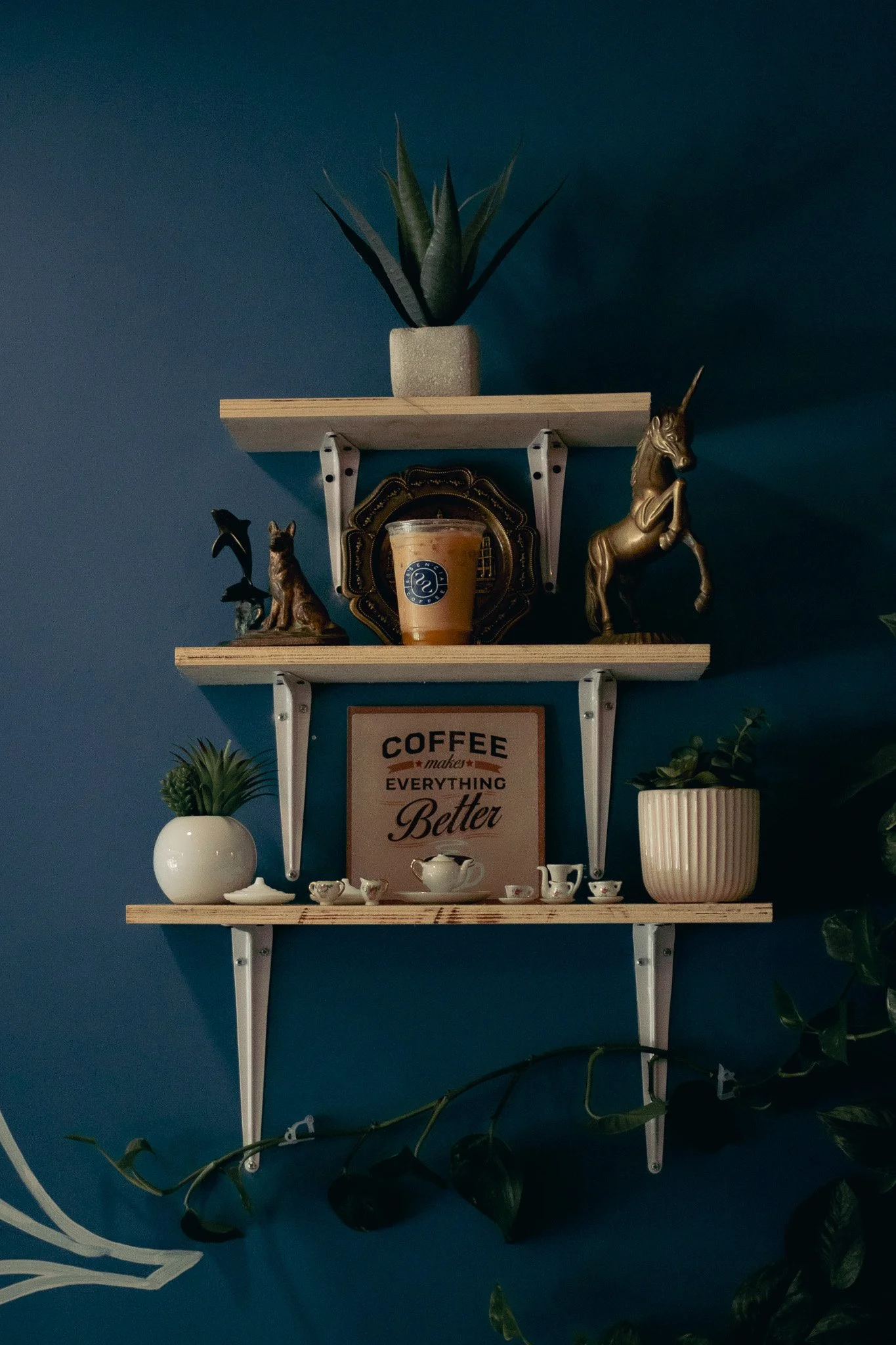 Estantes de madera con decoraciones en una pared azul, incluyendo plantas, figurinas de animales y un cartel que dice 'Coffee makes everything better', con una bebida en vaso de café en la parte media.