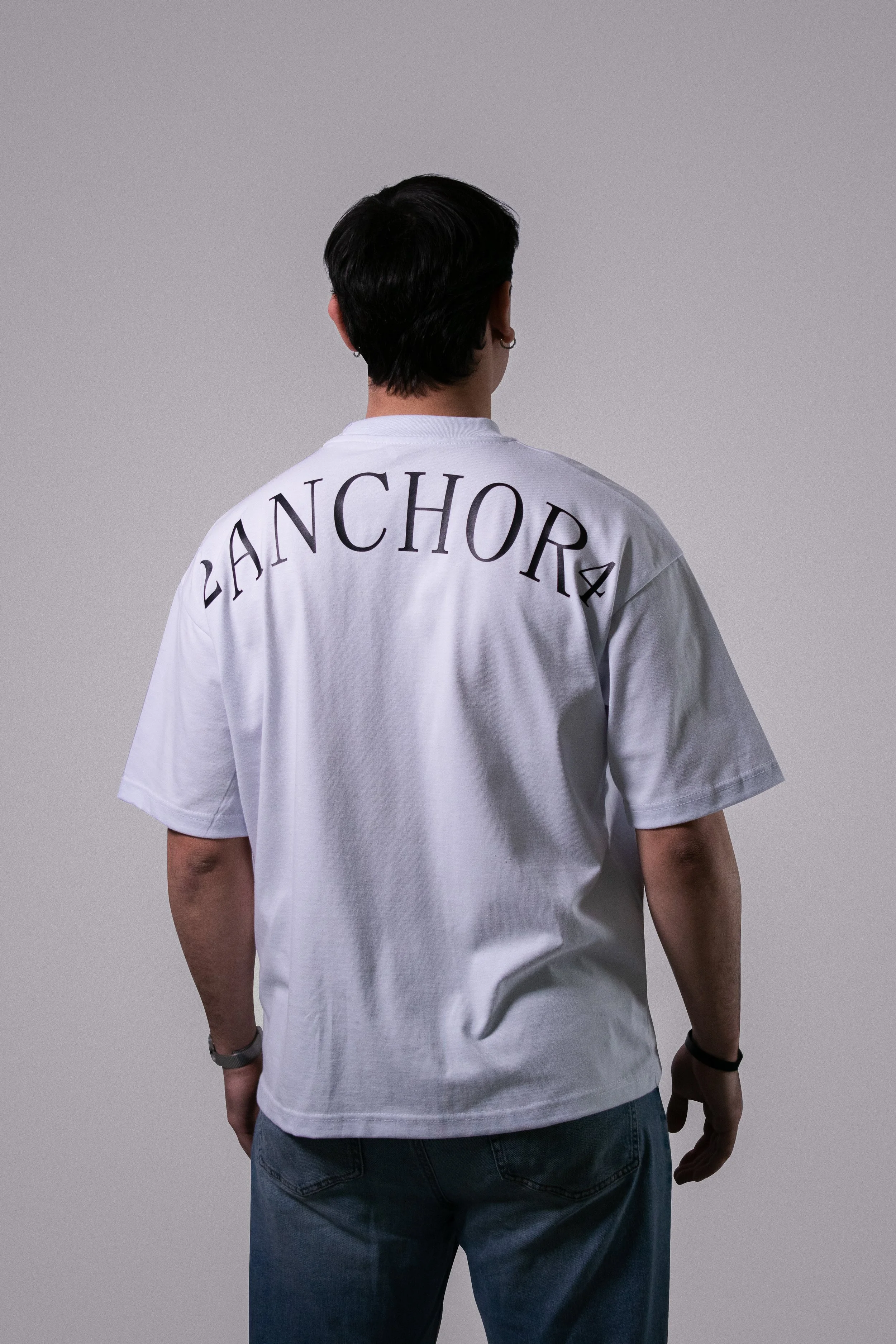 Persona vista desde atrás con camiseta blanca que tiene la palabra 'ANCHOR' escrita en letras grandes en la parte superior, cabello oscuro, y usando pulsera en la muñeca derecha.