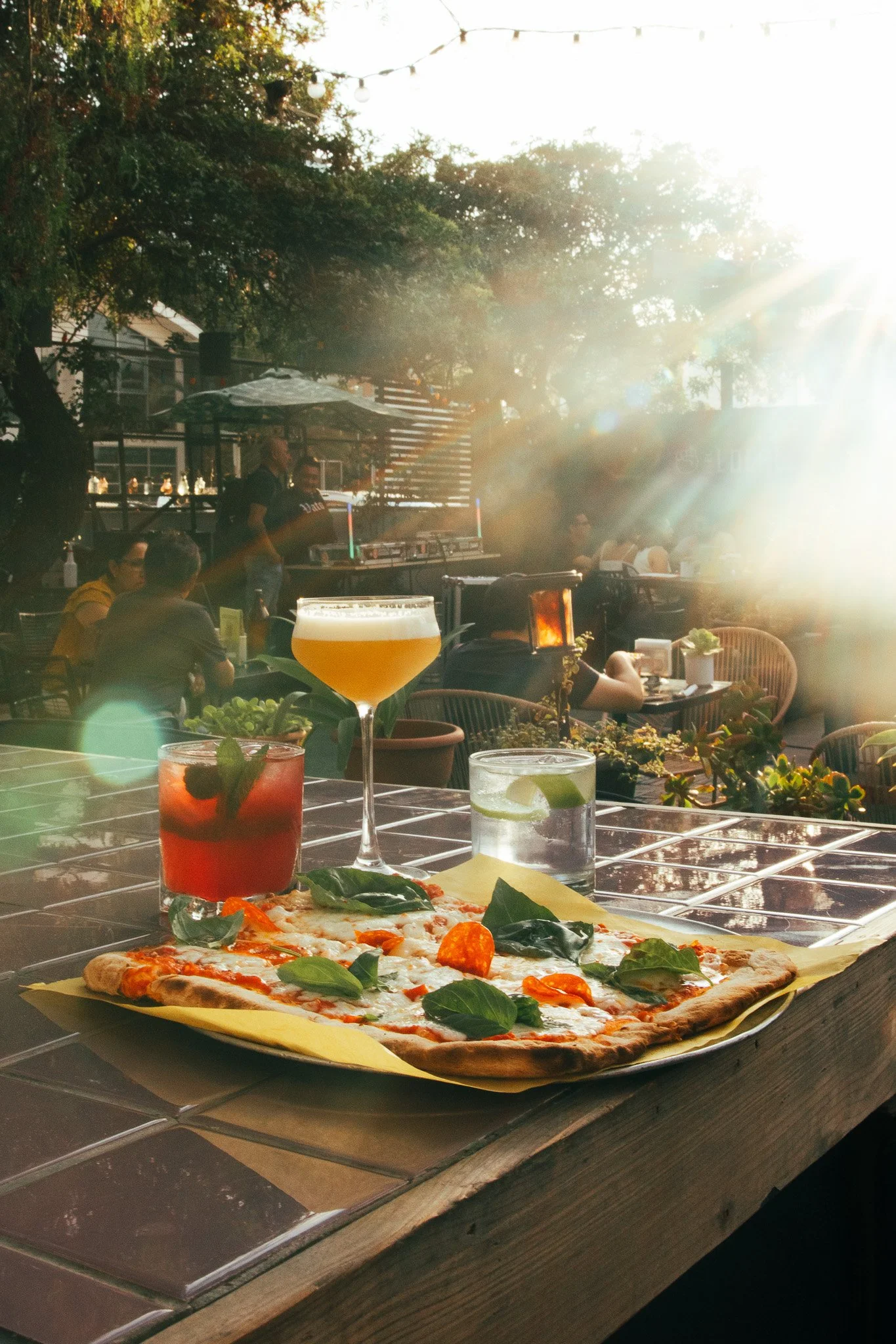 Pizza margarita con hojas de albahaca, limonada y cócteles en una mesa de madera en un restaurante al aire libre con luz solar brillante y gente sentada en el fondo.