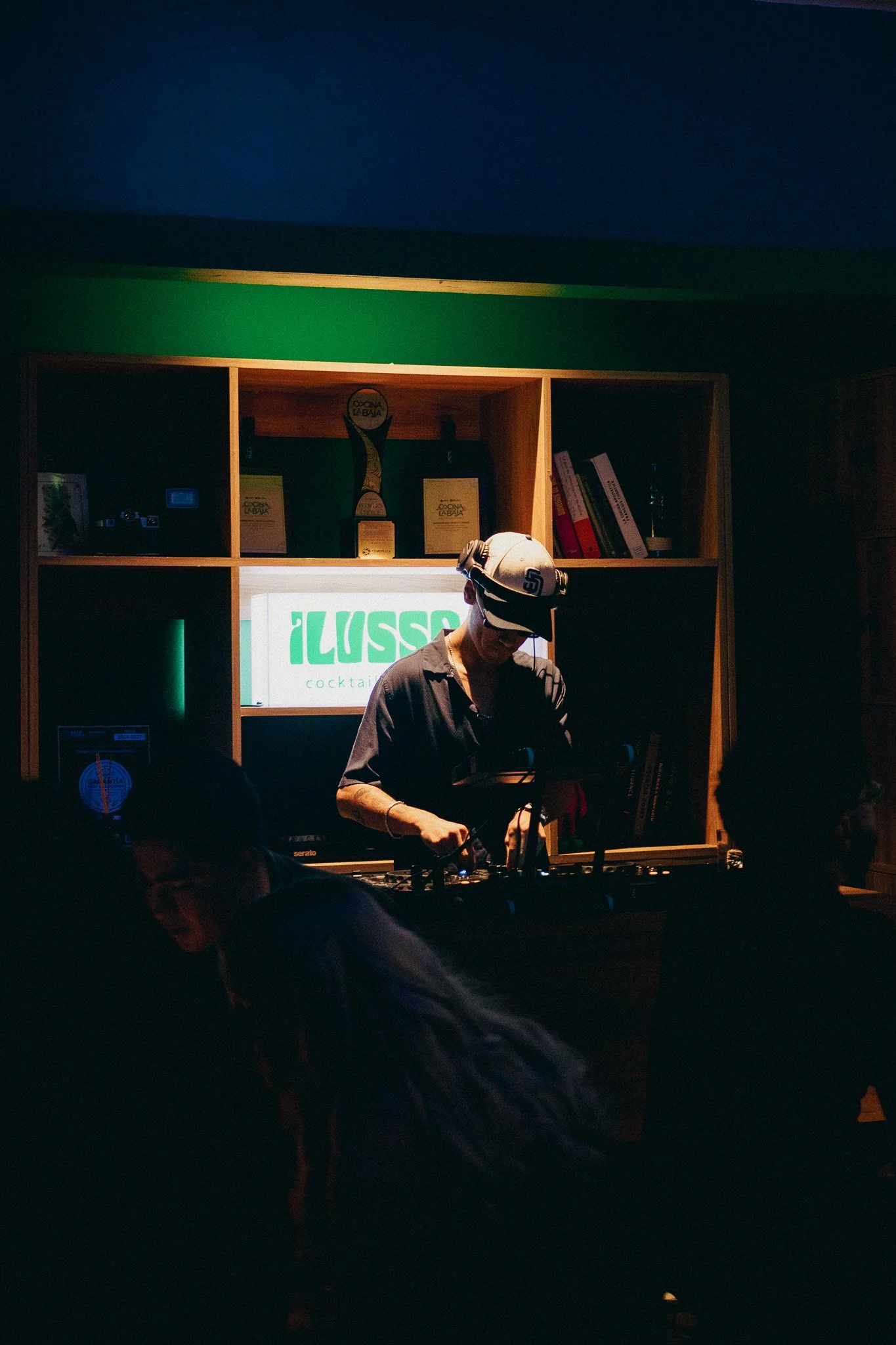 Un DJ tocando en un establecimiento llamado 'ILUSSI cocktail' con una estantería de madera atrás, en un ambiente oscuro y con algunas personas en primer plano.