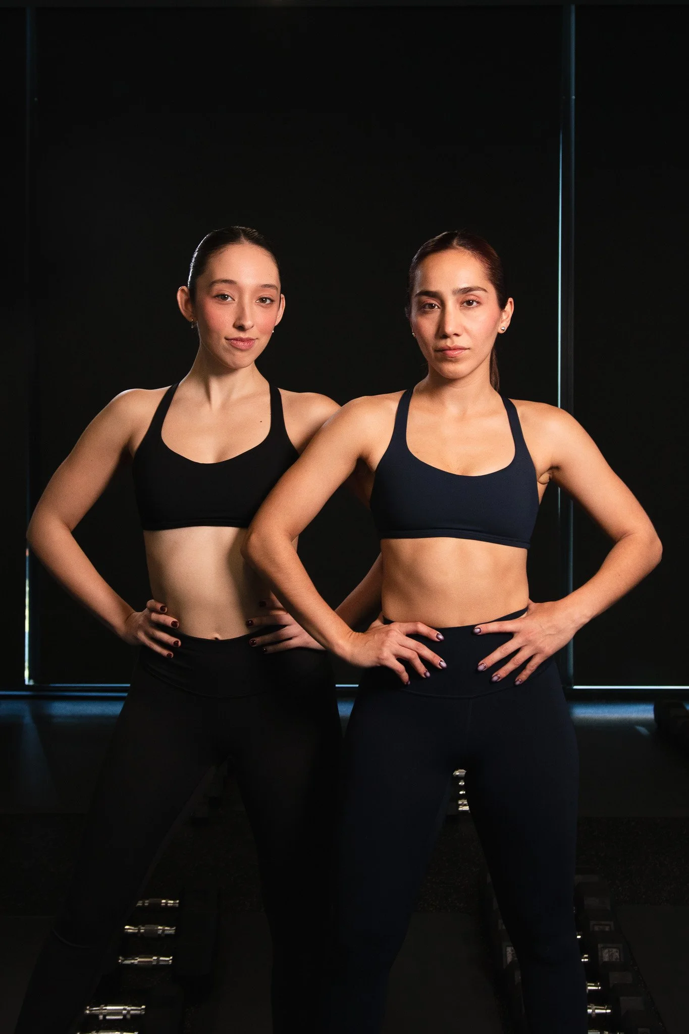 Dos mujeres en ropa de entrenamiento de yoga o fitness, de pie y con las manos en las caderas, en un ambiente oscuro con fondo negro.