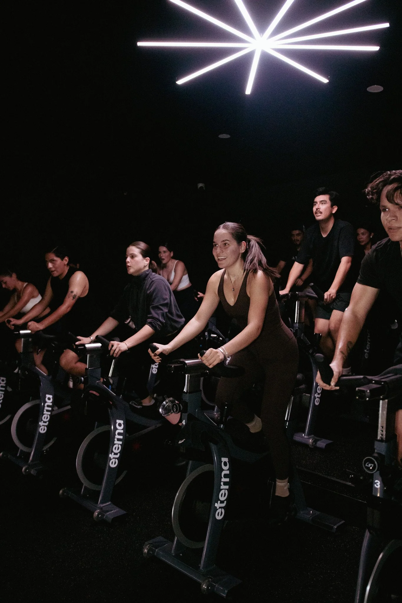 Grupo de personas participando en una clase de cycling en un estudio oscuro con iluminación moderna en el techo.