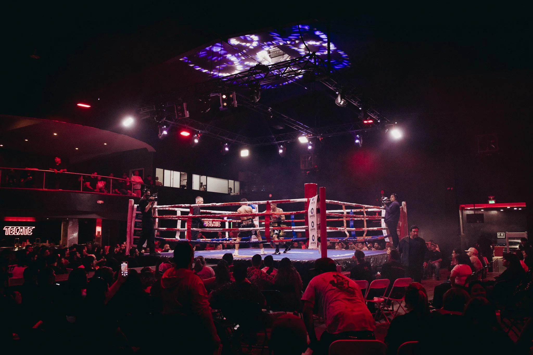 Una pelea de boxeo en un ring con espectadores en un evento nocturno, con luces brillantes y cámaras filmando.