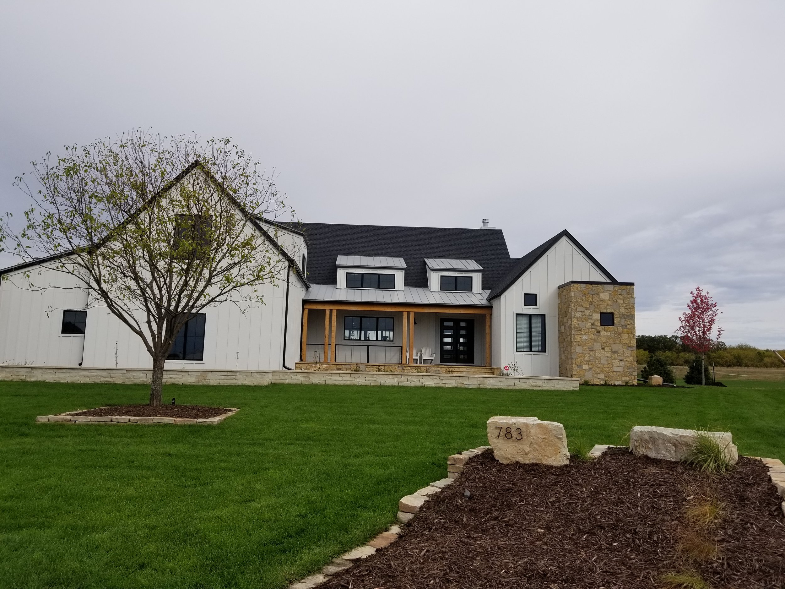 Modern house with white siding, a stone accent wall, and a front porch with wooden columns, surrounded by a green lawn with trees and a garden bed.