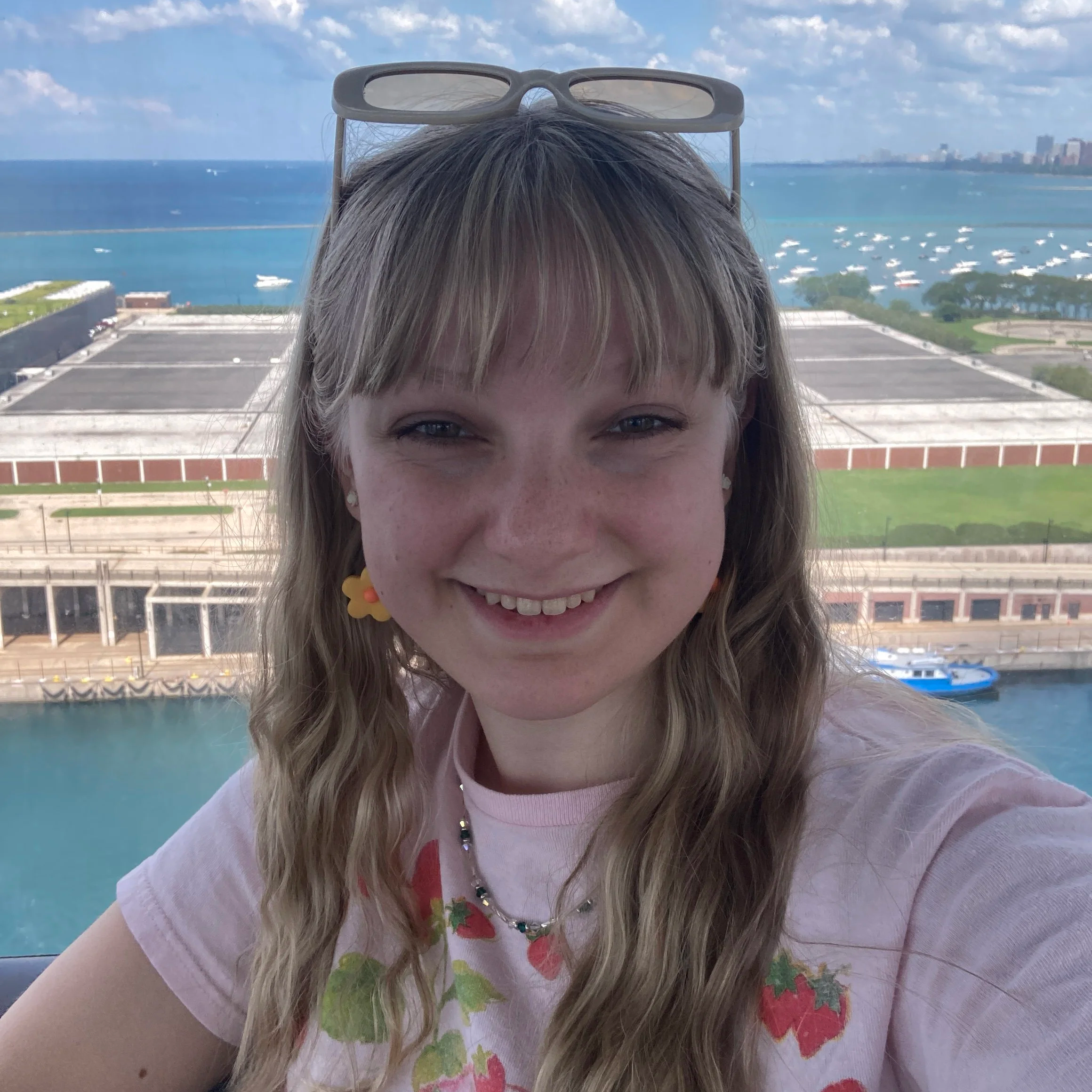An image of Lucia Sammarco at Navy Pier. They are a feminine-presenting person with wavy blonde hair, bangs, and green eyes. She is wearing a pink shirt, orange flower earrings, and beige sunglasses.