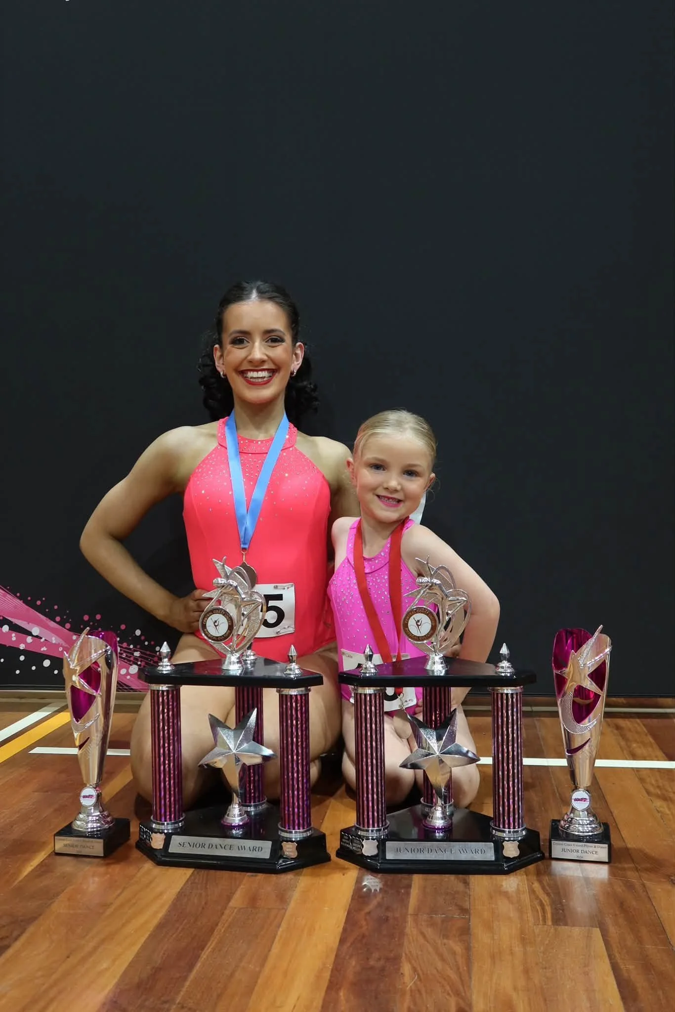 Two young female dancers in competition leotards standing on a gymnastics floor holding trophies, smiling, with a backdrop that reads 'CCUPD Central Coast United Physie & Dance'.