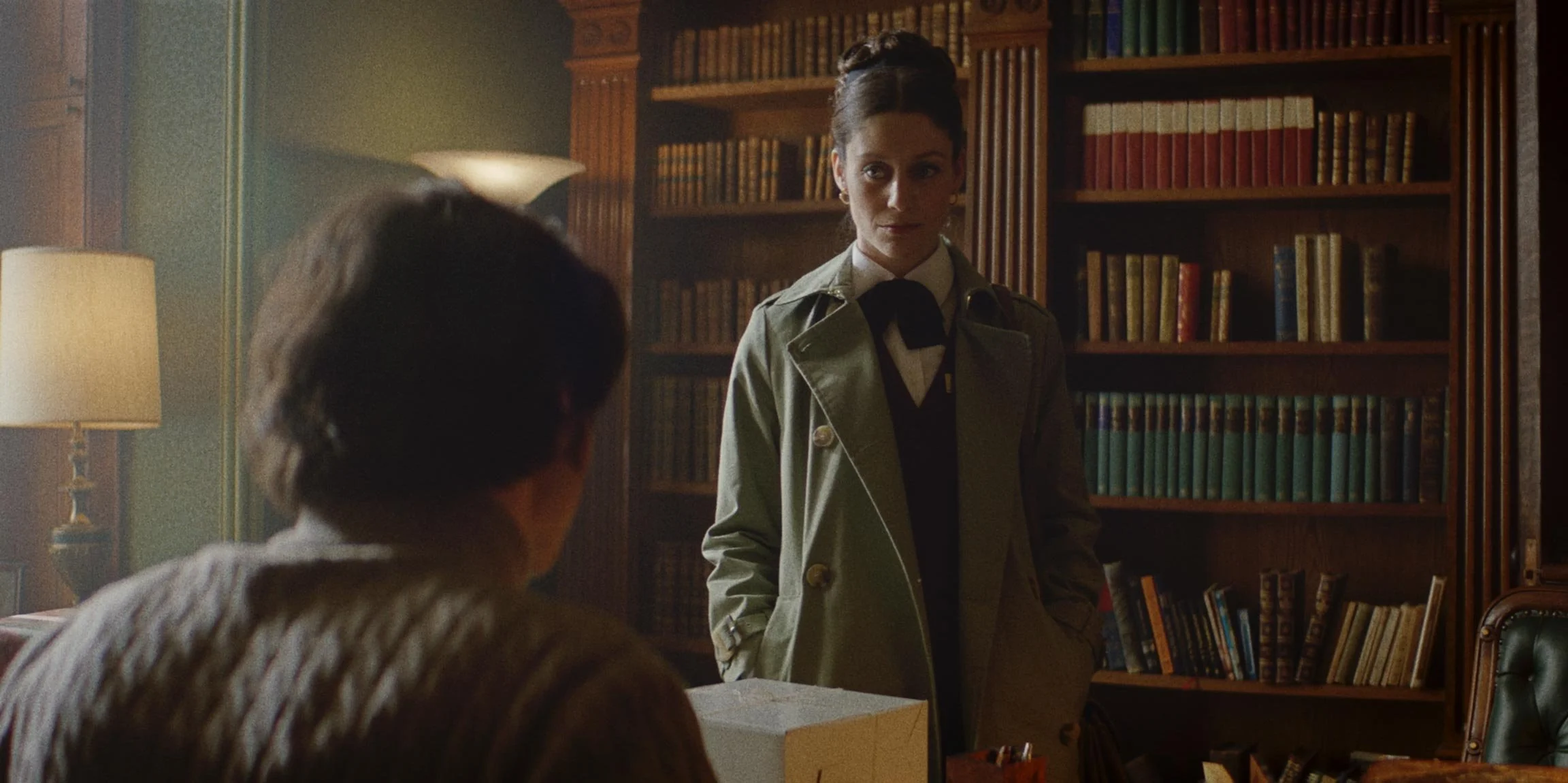 A woman standing in a room with wooden bookshelves, facing an older man sitting at a desk, with a table lamp illuminating the scene.